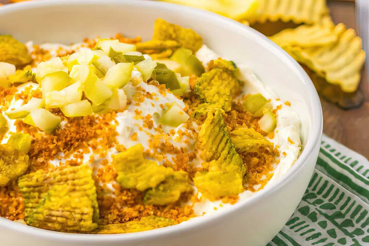 The image features a bowl of dip topped with crushed chips and diced pickles, surrounded by crinkle-cut chips.