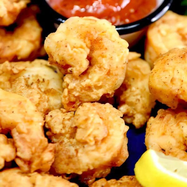 Crispy fried shrimp appetizers with lemon wedges and a container of red dipping sauce in the background.