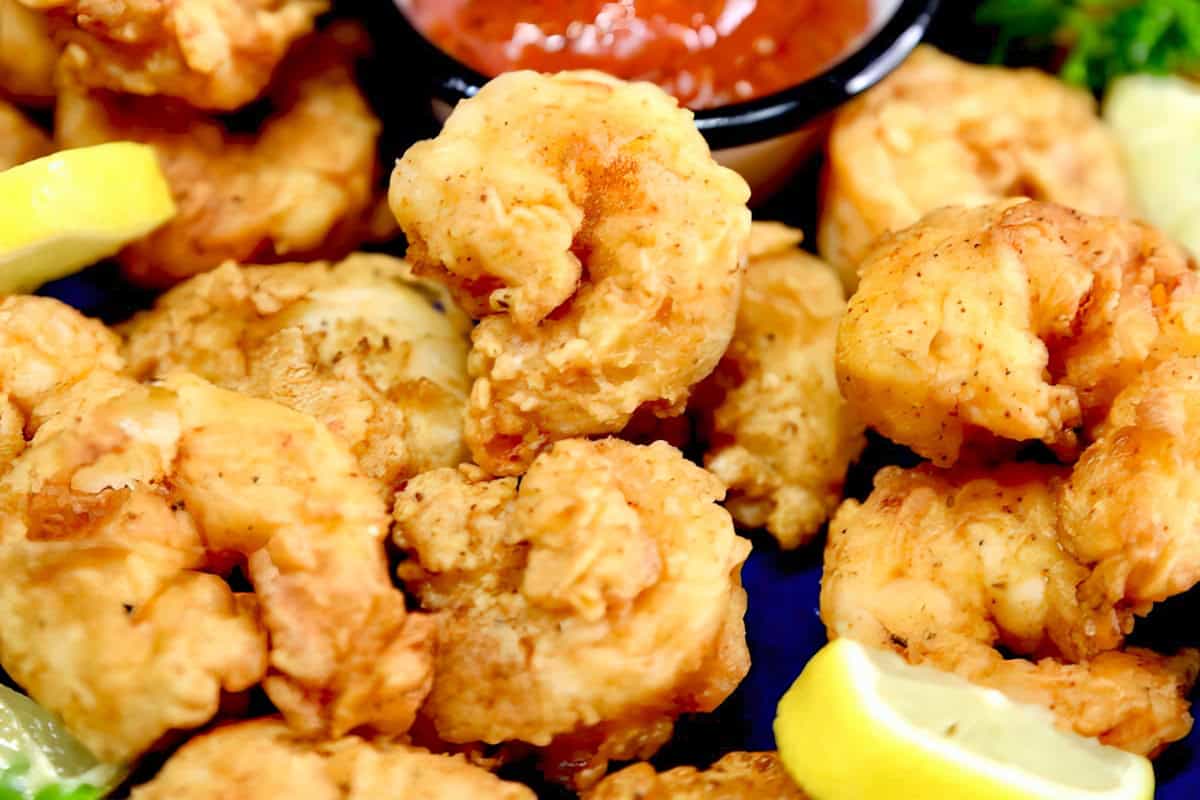 Close-up of fried shrimp served with lemon wedges and a cup of red dipping sauce in the background. The shrimp are golden brown and crispy, suggesting a freshly cooked, appetizing dish.