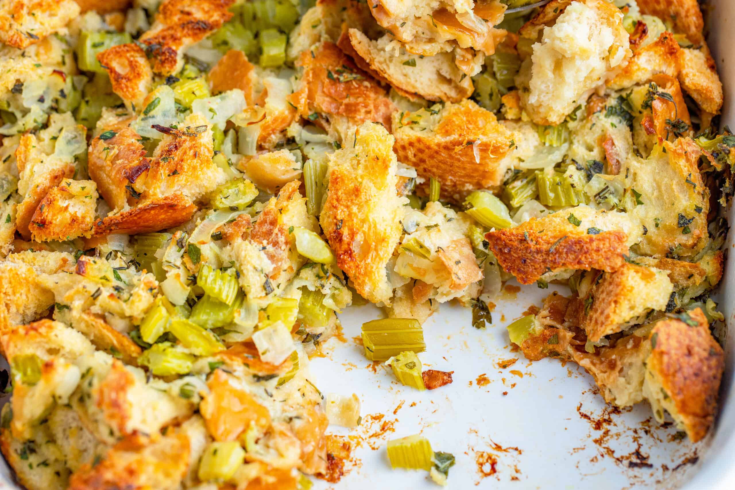 The image shows a pan filled with baked stuffing, featuring crispy bread pieces mixed with chopped celery and herbs.