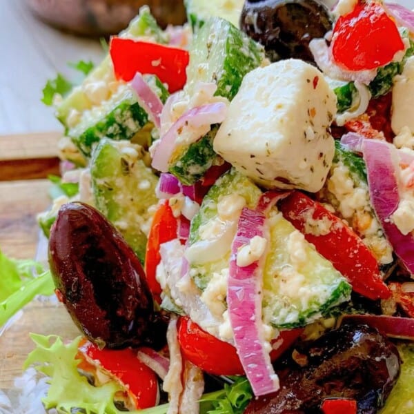 The image displays ripe tomatoes, crisp cucumbers, red onions, creamy feta cheese, kalamata olives, and fresh lettuce arranged on a rustic wooden surface.