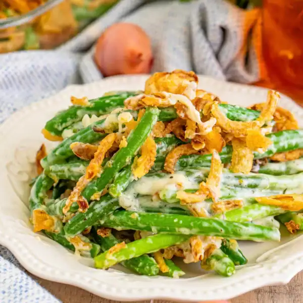 A serving of green bean casserole topped with crispy fried onions on a white plate, with a drink and more casserole in the background.