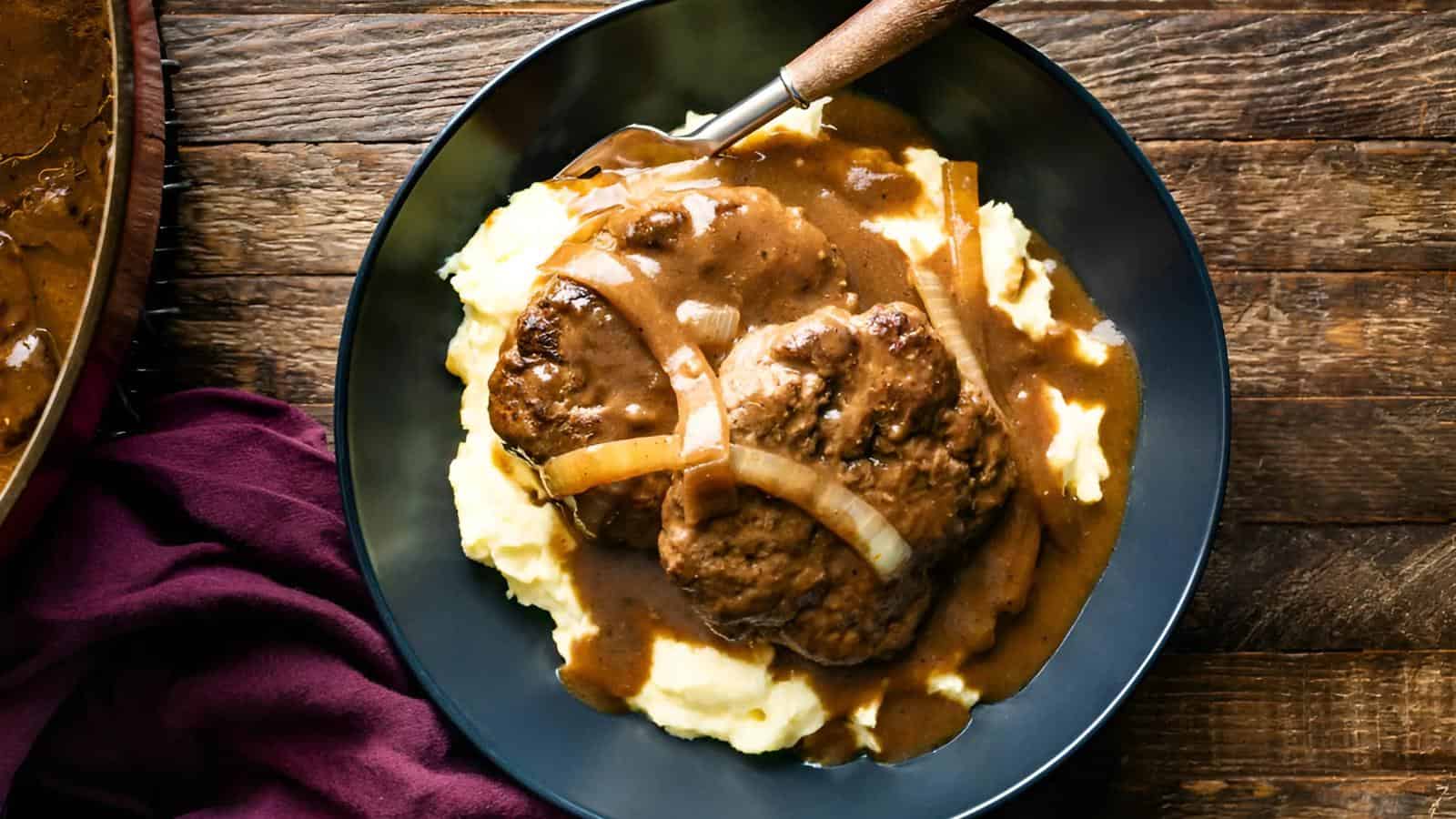 A black plate featuring mashed potatoes topped with two beef patties, gravy, and onion slices.