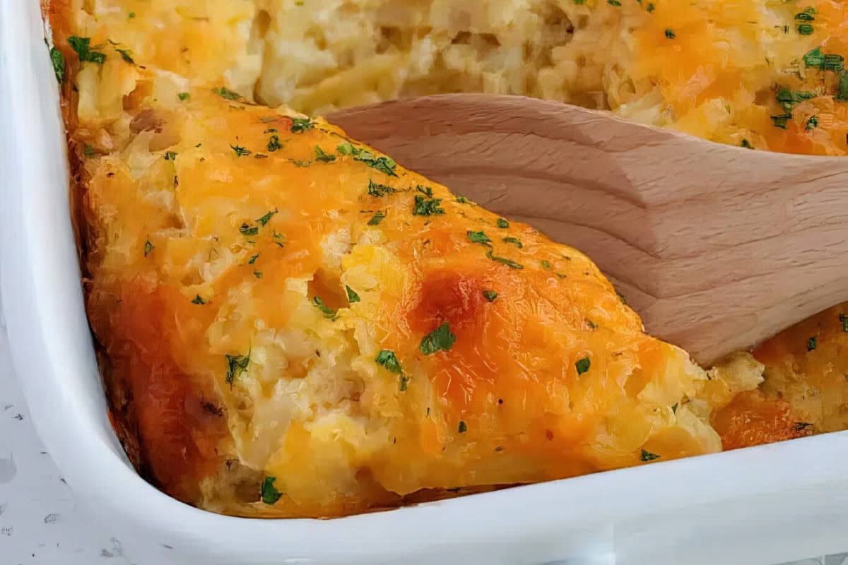 A baked potato casserole with a golden crust in a white dish, accompanied by a wooden serving spoon.