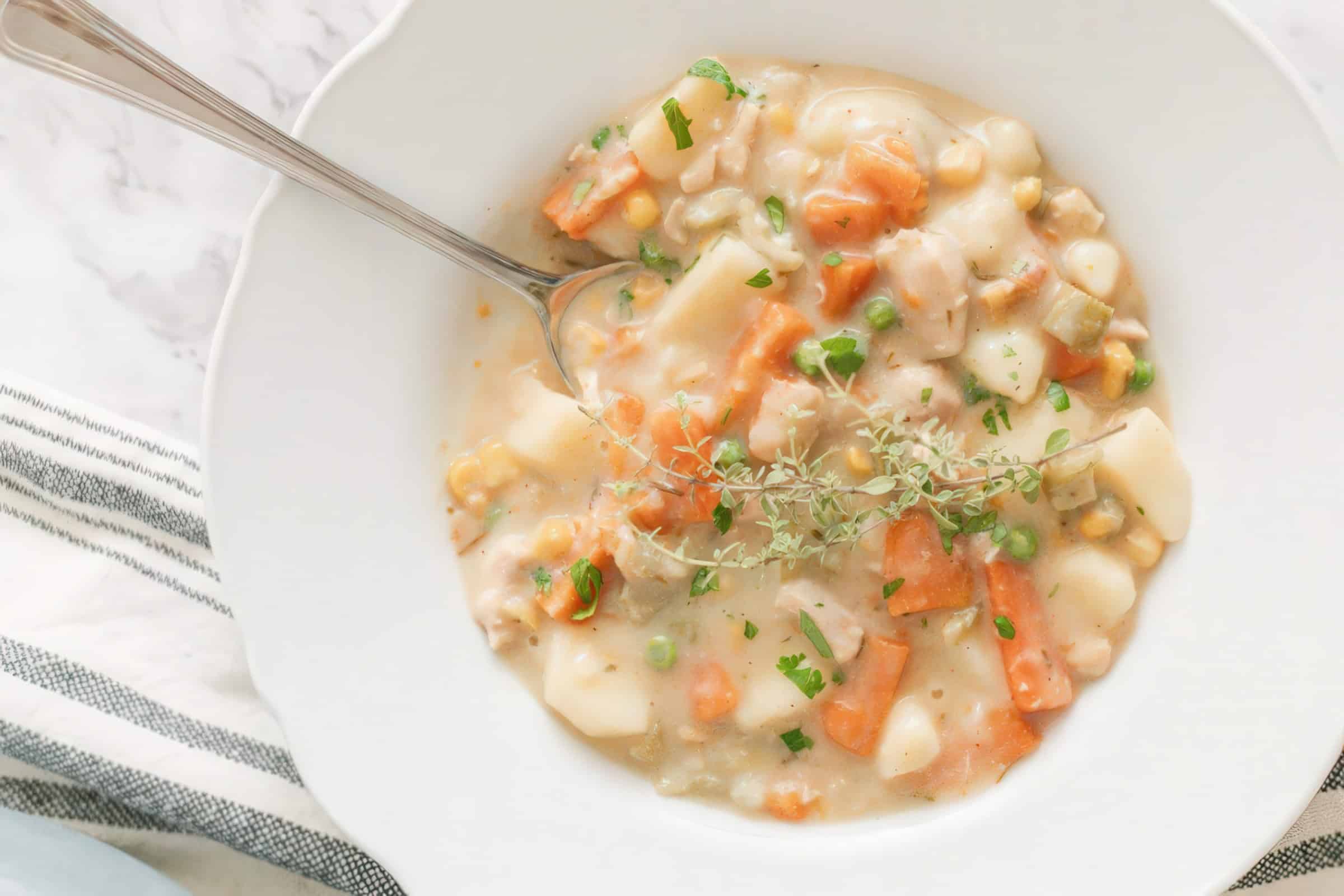 A bowl of creamy vegetable soup featuring carrots, potatoes, corn, peas, and herbs, with a spoon resting in the bowl. The soup is garnished with herbs, and the table is set with a striped cloth.