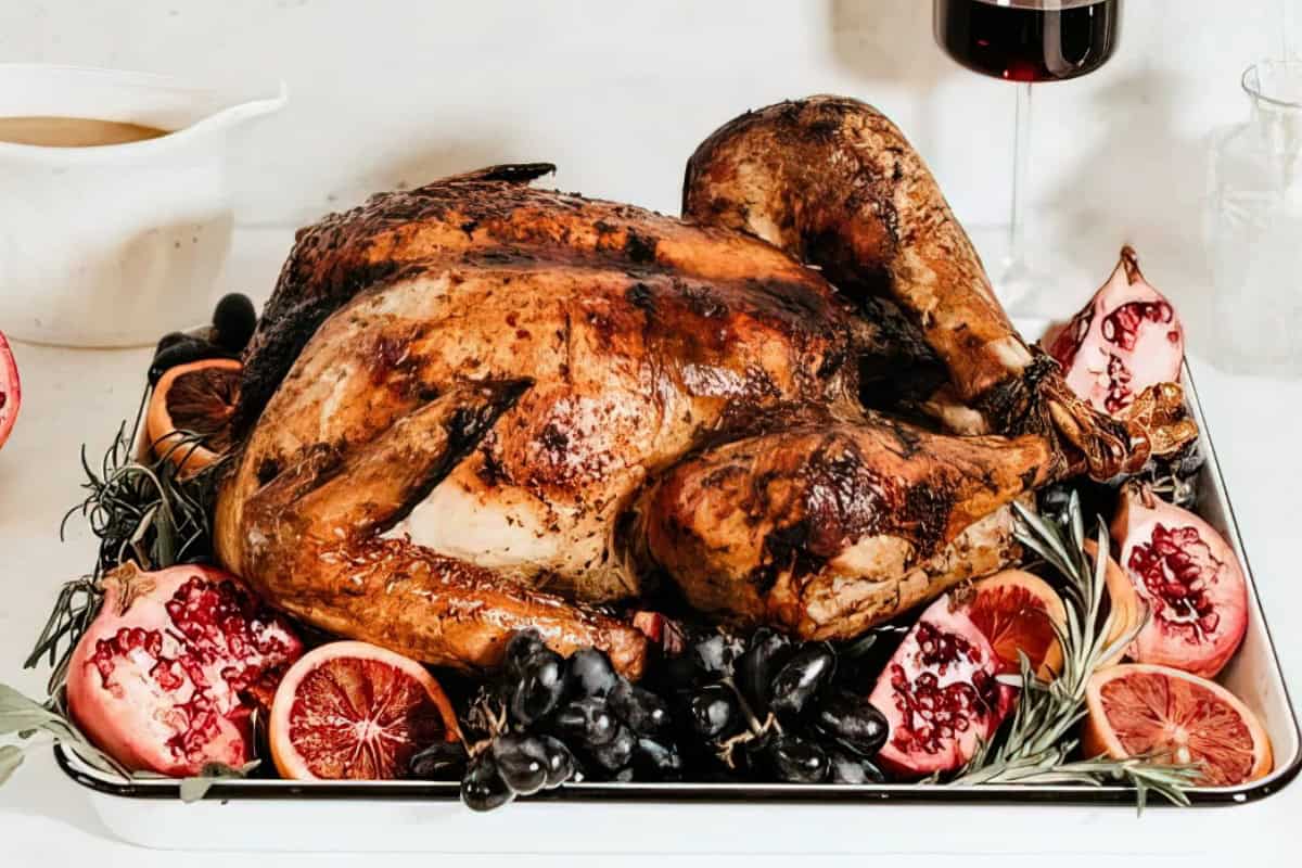 A roasted turkey on a platter, surrounded by sliced pomegranates, black grapes, and sprigs of rosemary. There's a white gravy boat on the left and a glass of red wine in the background.