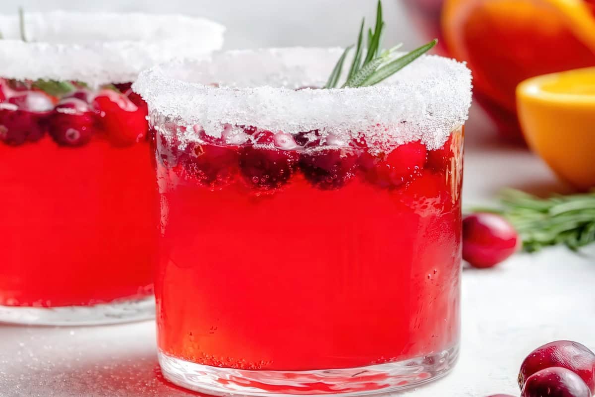 A close-up of two glasses filled with a vibrant red cocktail. The rims are sugar-coated, and each drink is garnished with cranberries and a sprig of rosemary. There's a blurred orange slice in the background.