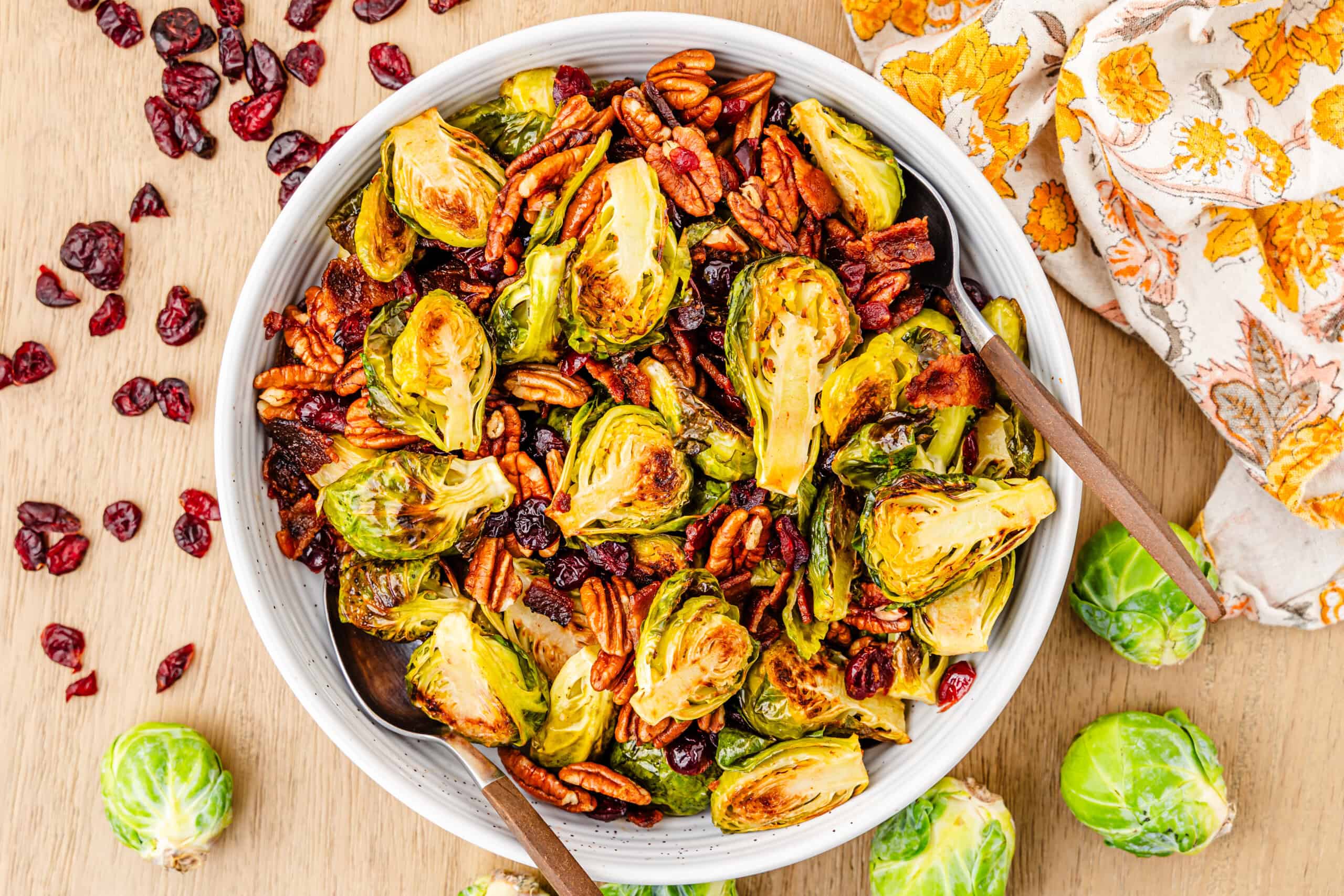 The image features a bowl containing roasted Brussels sprouts, pecans, and cranberries. Fresh Brussels sprouts and cranberries are scattered on a wooden table around the bowl. Inside the bowl are two wooden spoons.