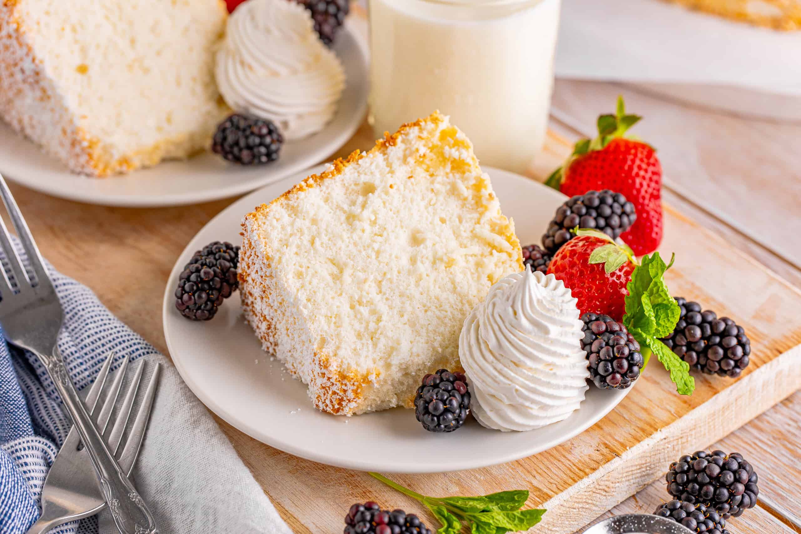 The image shows a slice of cake on a white plate, along with whipped cream, strawberries, and blackberries. Beside the plate is a glass of milk.