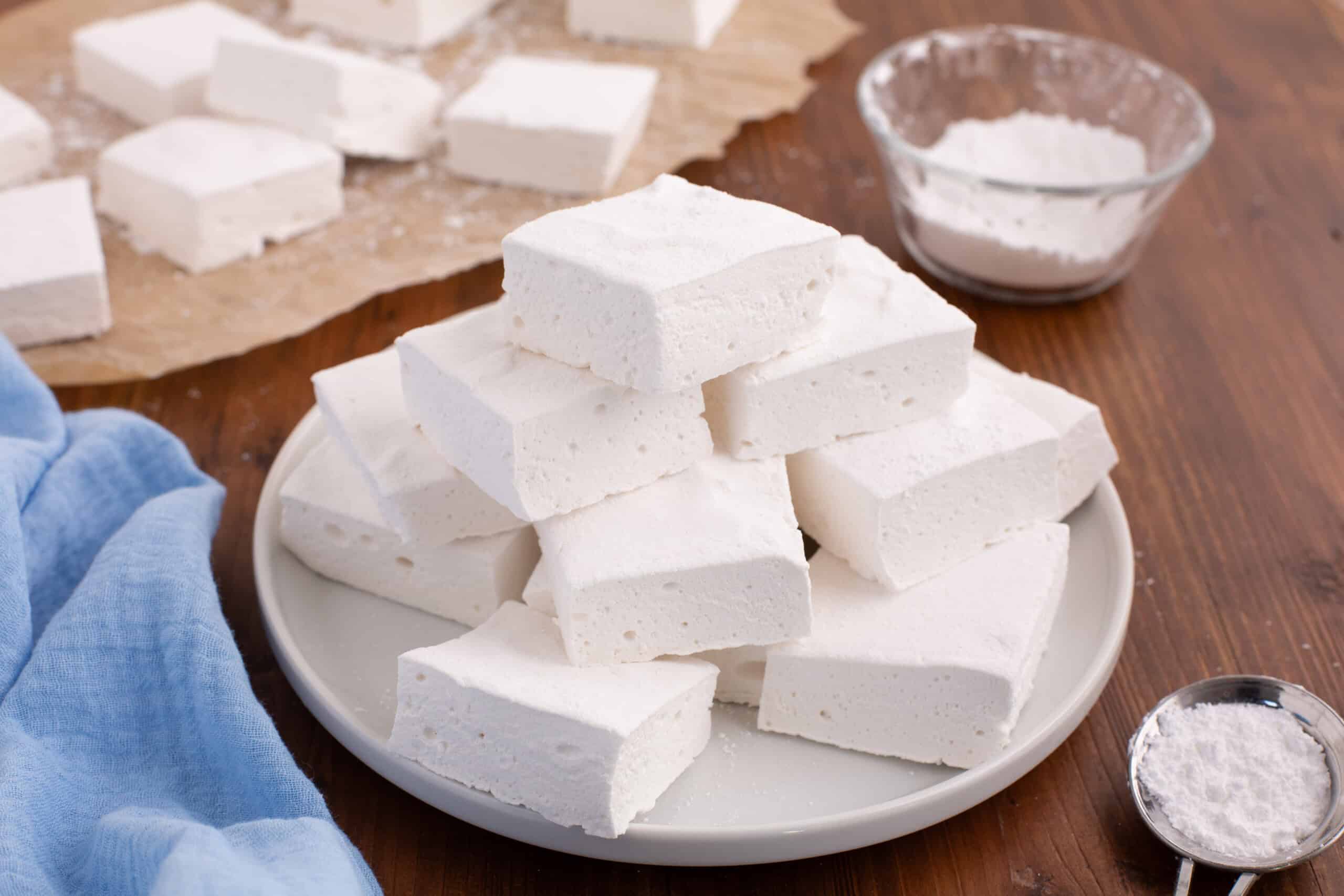 A stack of homemade marshmallows on a white plate, a bowl of powdered sugar, and a blue cloth.