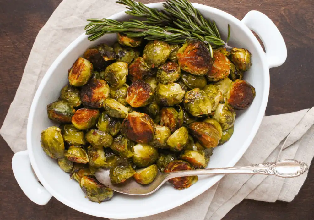 A white dish containing roasted Brussels sprouts with a sprig of rosemary, a silver serving spoon inside, placed on a wooden table and accompanied by a beige cloth.