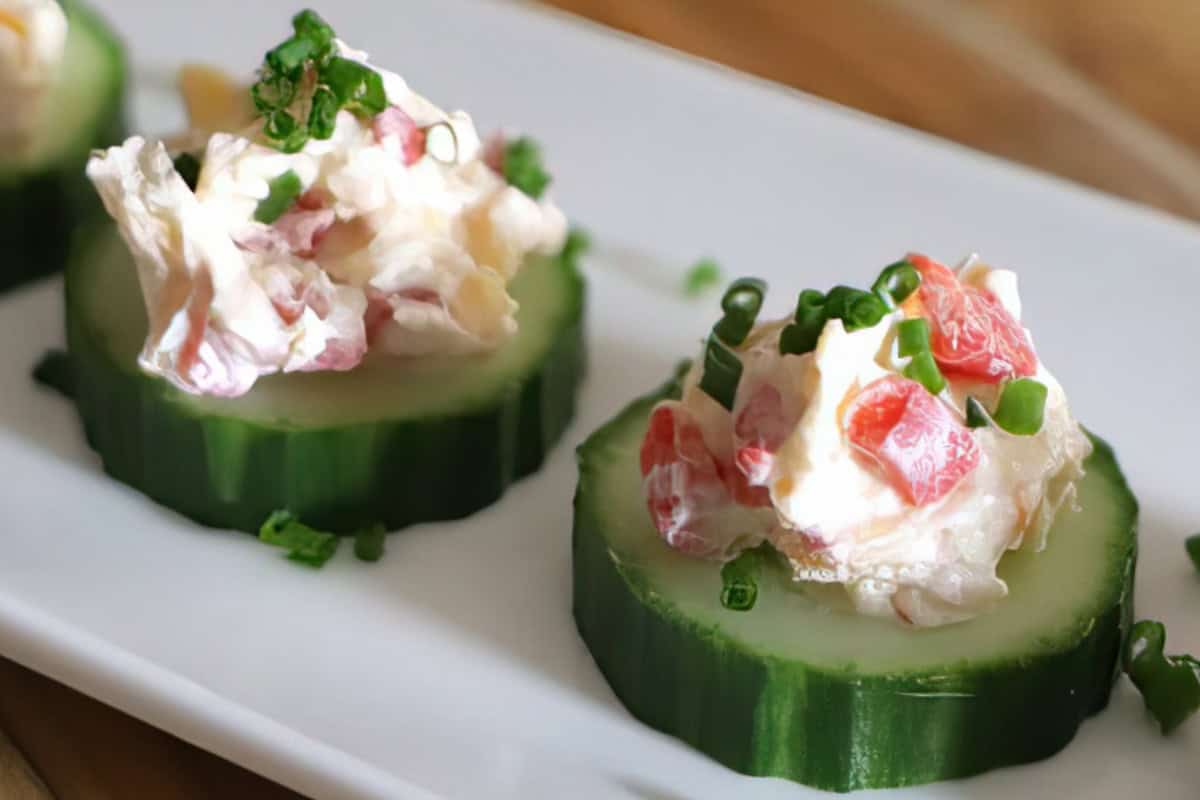 Cucumber slices topped with a creamy mixture of diced red bell peppers and garnished with chopped chives on a white plate.