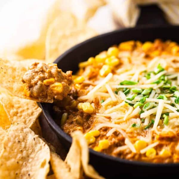 A skillet filled with cheesy dip containing ground meat and corn, topped with shredded cheese and green herbs, surrounded by tortilla chips.