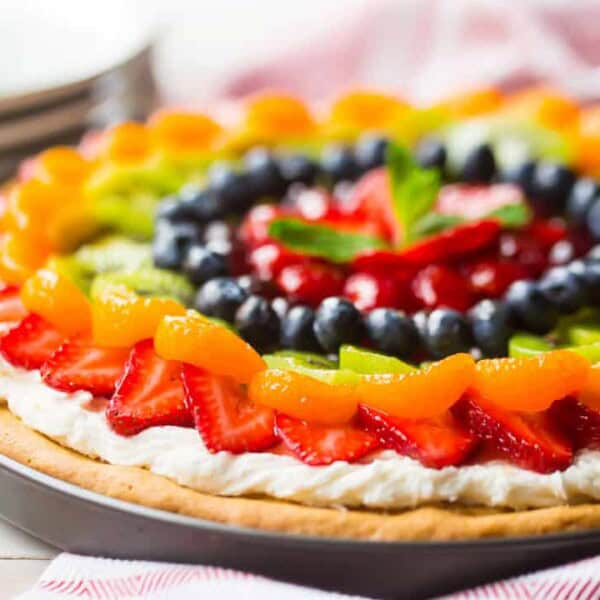 A fruit pizza on a round tray with a creamy base, topped with sliced strawberries, mandarin oranges, kiwi, blueberries, and grapes.