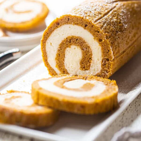 A sliced pumpkin roll cake with cream cheese filling sits on a rectangular white plate, with a pen and napkin close by.