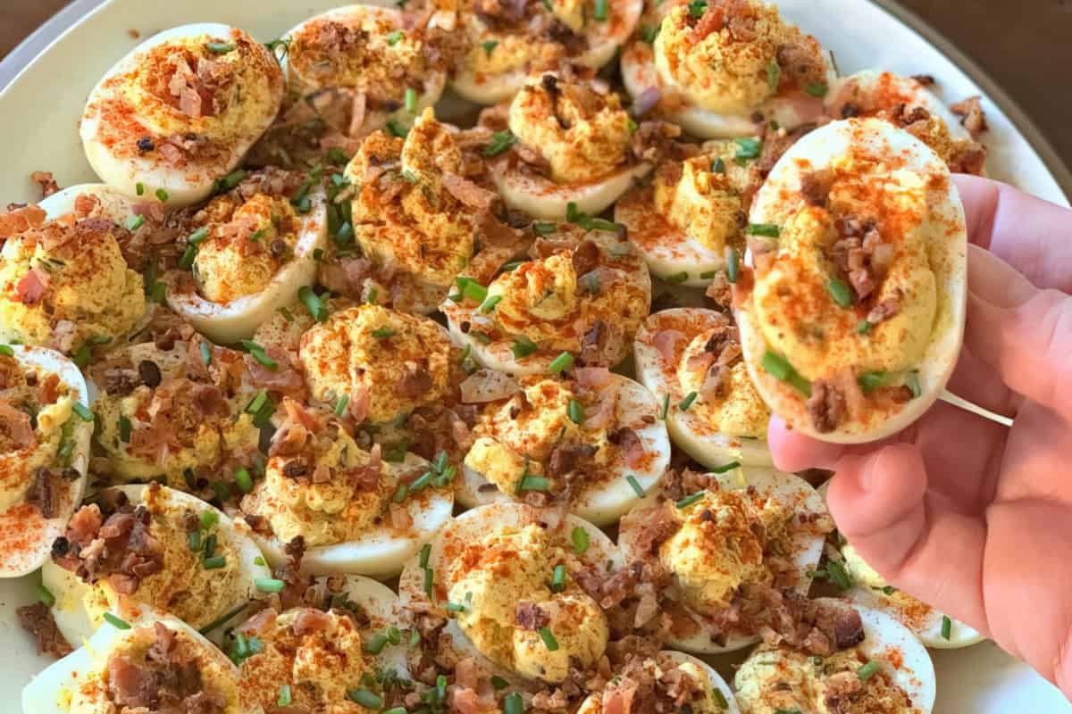 A plate of deviled eggs topped with bacon bits, chives, and paprika. A single deviled egg is held above the plate.