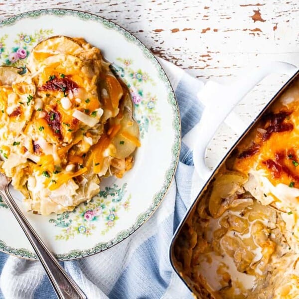 Cheesy scalloped potatoes on a floral plate with a fork, next to a baking dish containing the rest of the casserole.