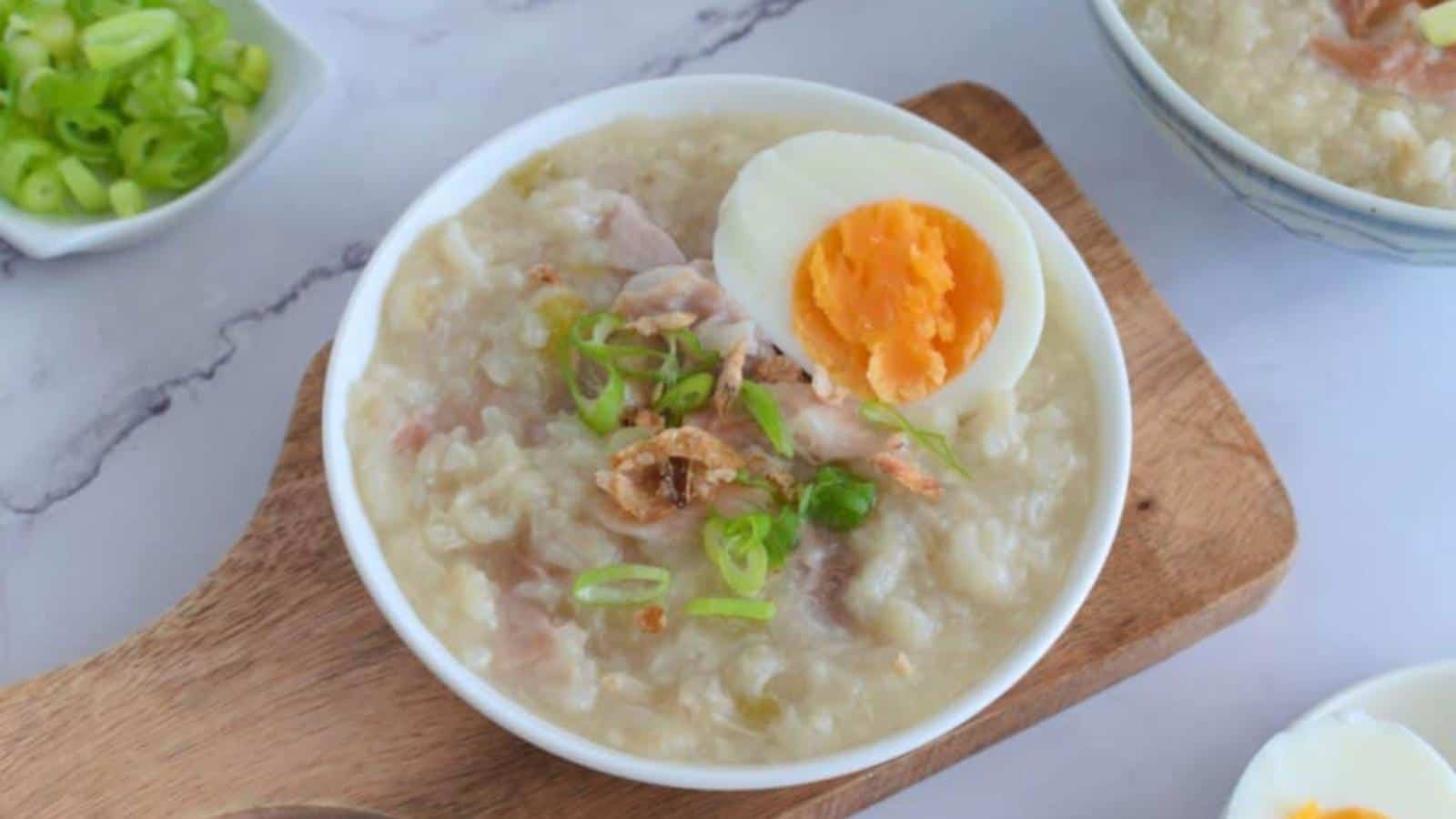 The image shows a bowl containing porridge topped with sliced green onions, shredded meat, and a halved boiled egg, all situated on a wooden tray.