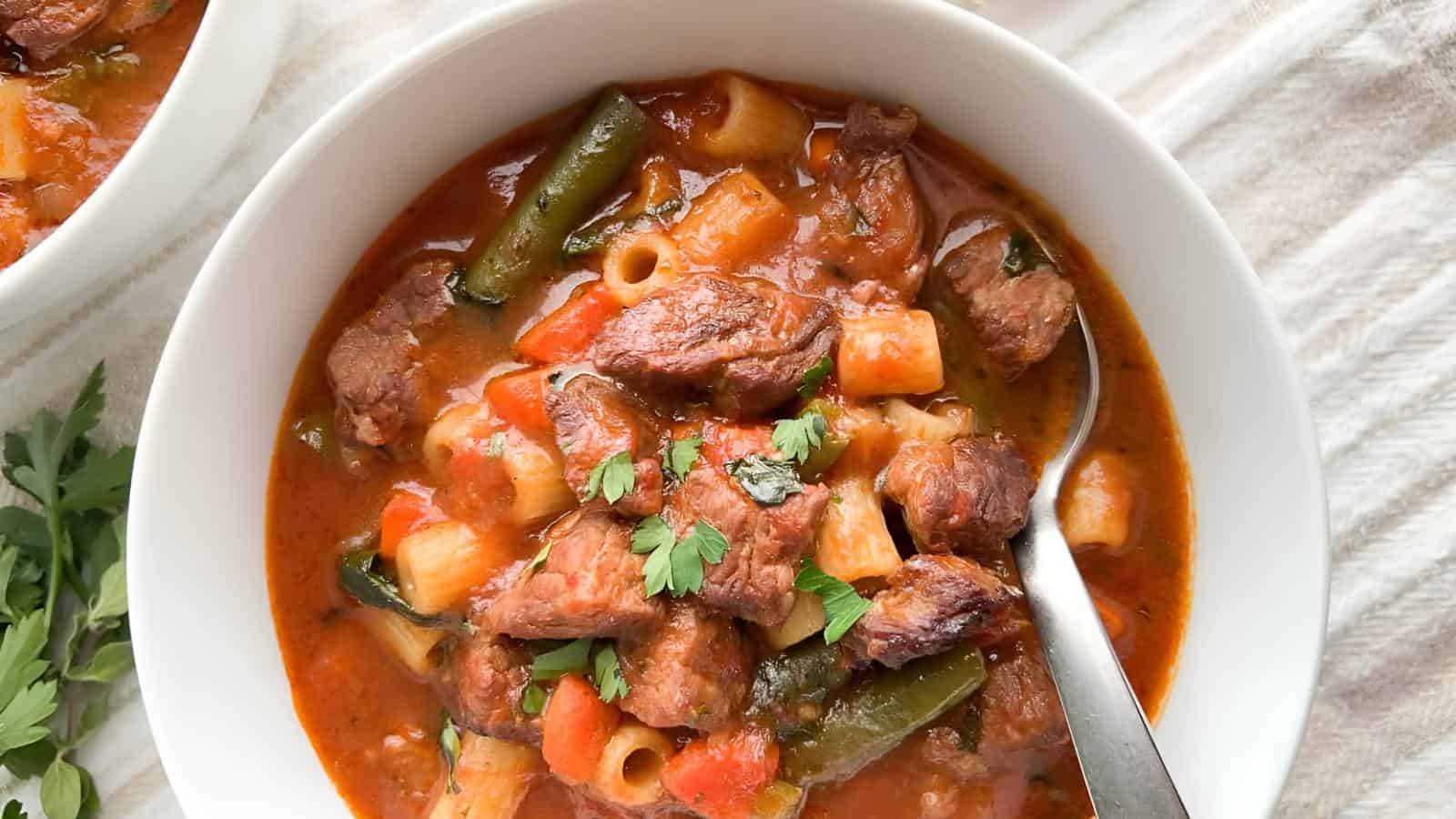 A bowl of hearty beef and vegetable soup with pasta, featuring chunks of beef, green beans, potatoes, and carrots in a rich broth. Garnished with fresh parsley, a spoon rests inside the bowl.