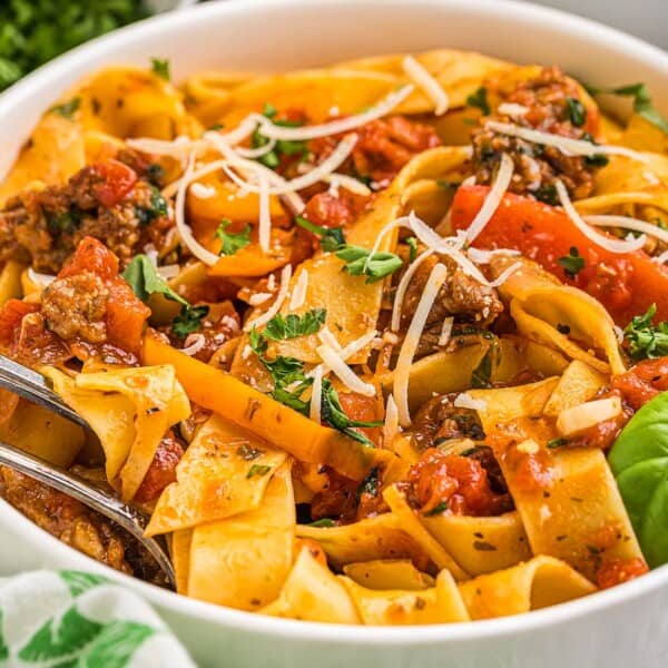 A bowl of pappardelle pasta with tomato sauce, ground meat, grated cheese, fresh herbs, and a fork and spoon for serving.