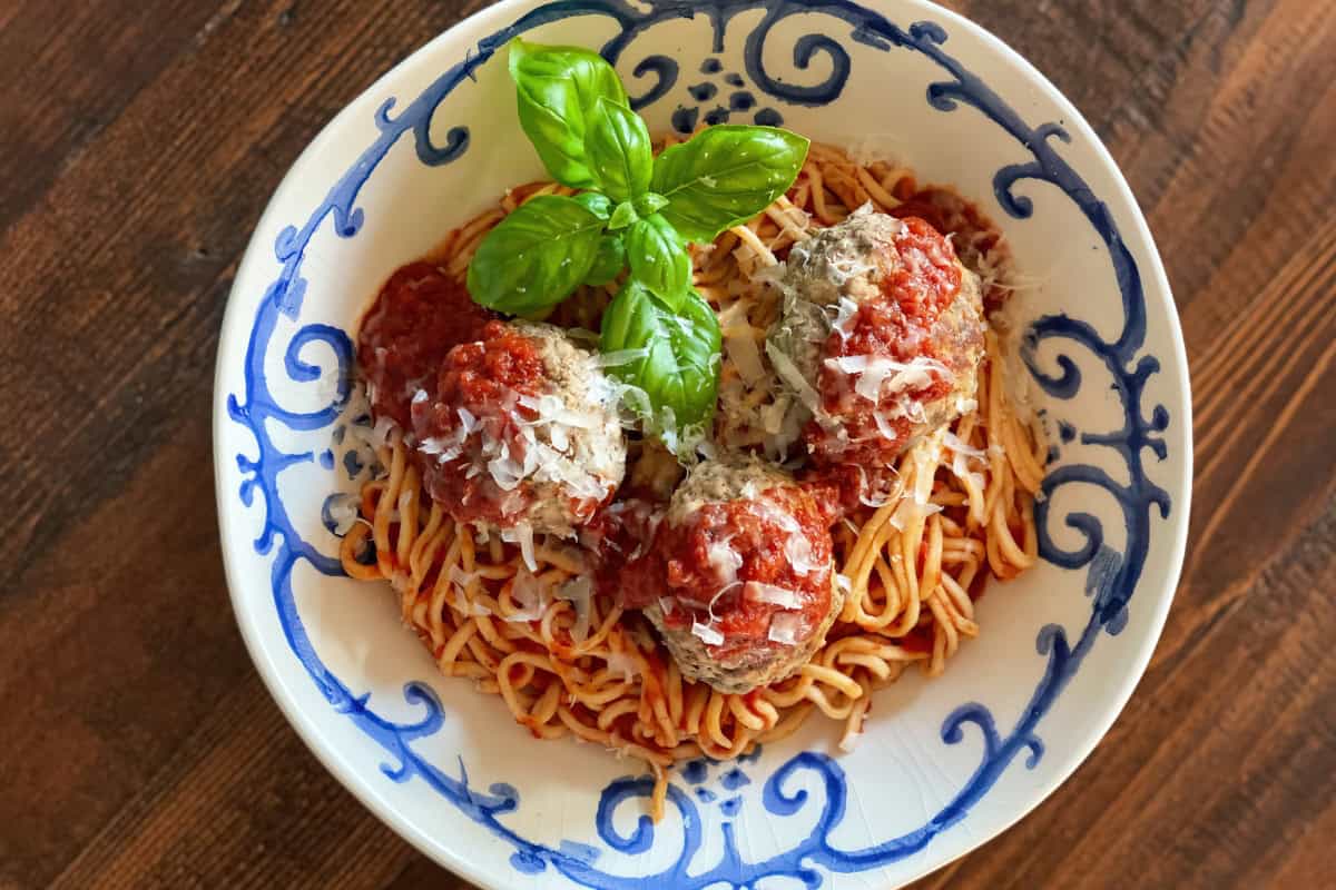 A decorative white and blue bowl on a wooden table, containing spaghetti with three meatballs, tomato sauce, grated cheese, and a sprig of fresh basil.
