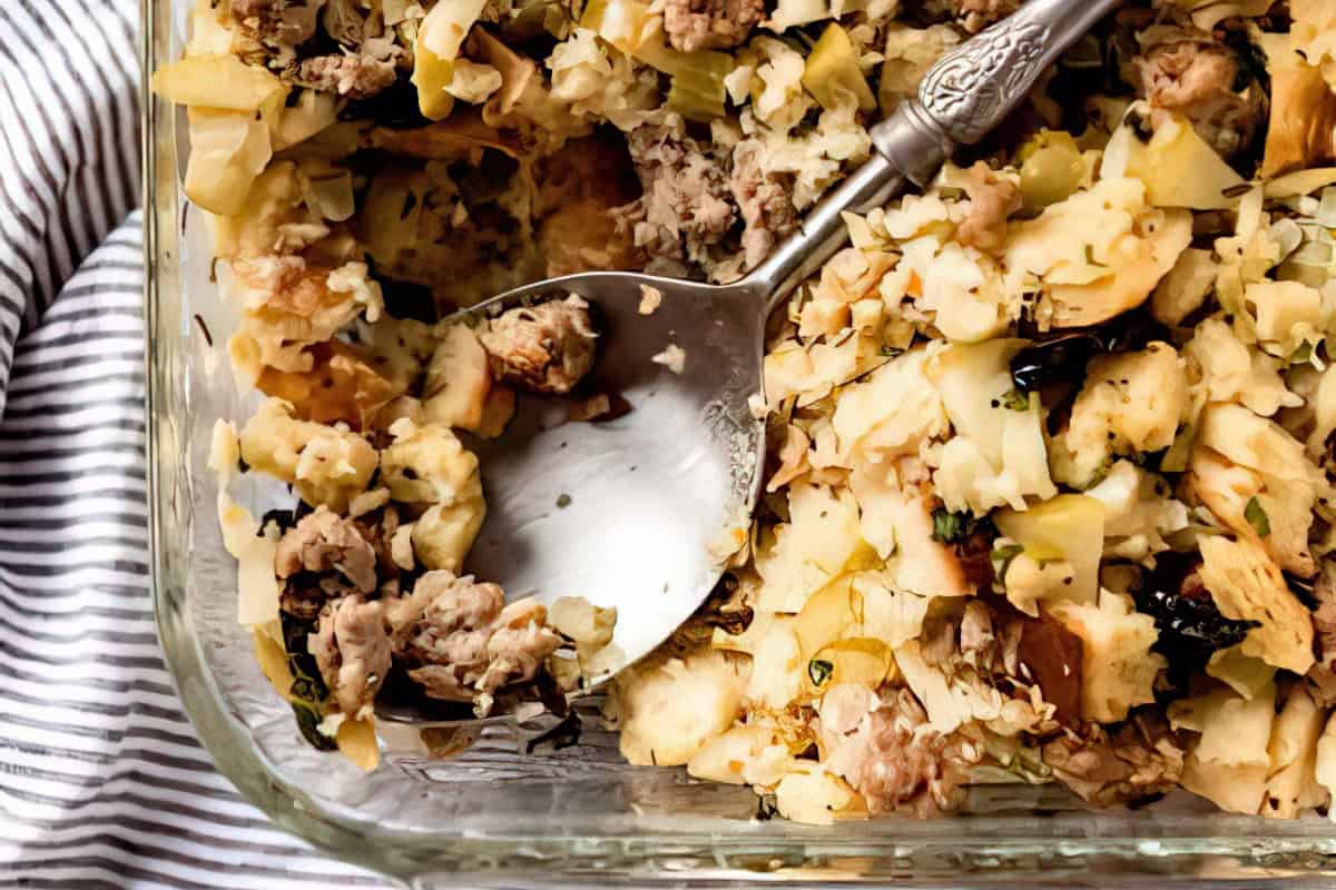 A close-up of a casserole dish filled with stuffing. The mixture includes chunks of bread, sausage, apples, and herbs. A silver serving spoon is resting in the dish. The dish is set on a striped cloth.