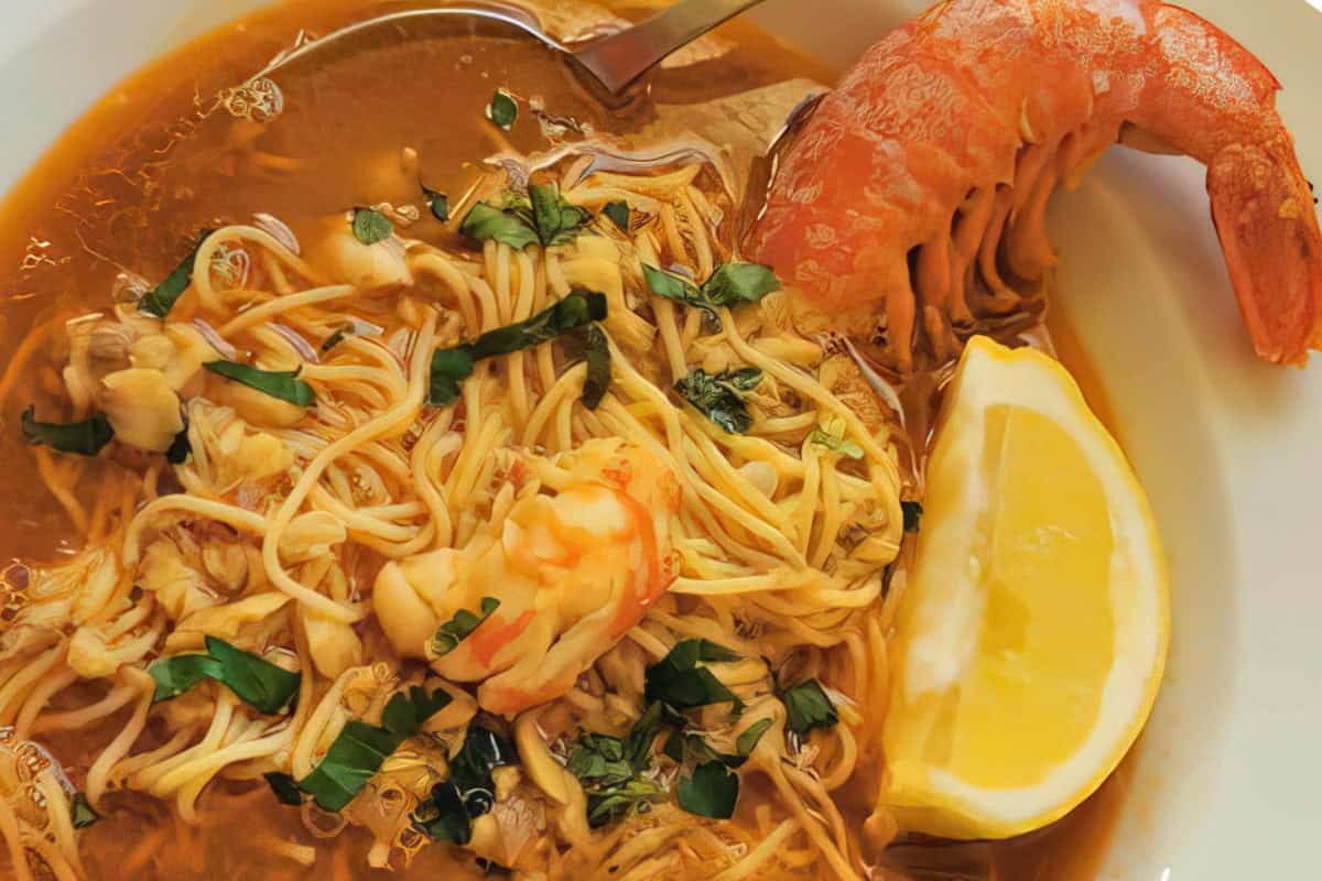 A bowl of soup featuring noodles, shrimp, and herbs, garnished with a lemon wedge. The soup has a rich broth, and a silver spoon rests in it alongside a large shrimp.