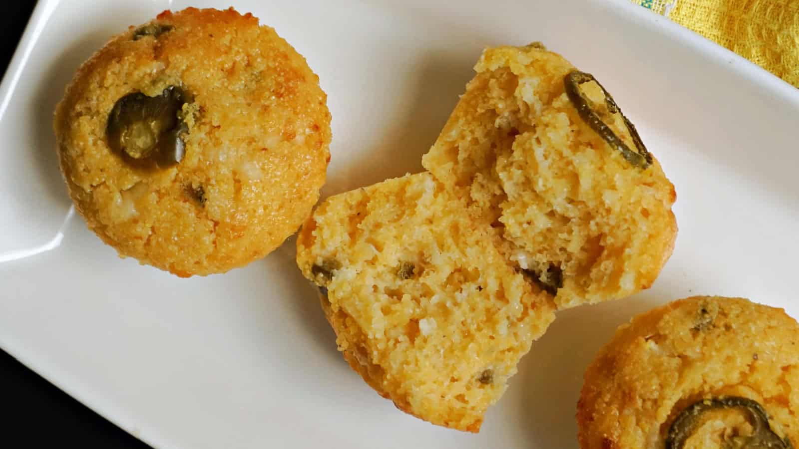 Three jalapeño cornbread muffins are on a white rectangular plate. One muffin is split in half, showing its texture and jalapeño pieces inside. The surface is golden brown with visible jalapeño slices.