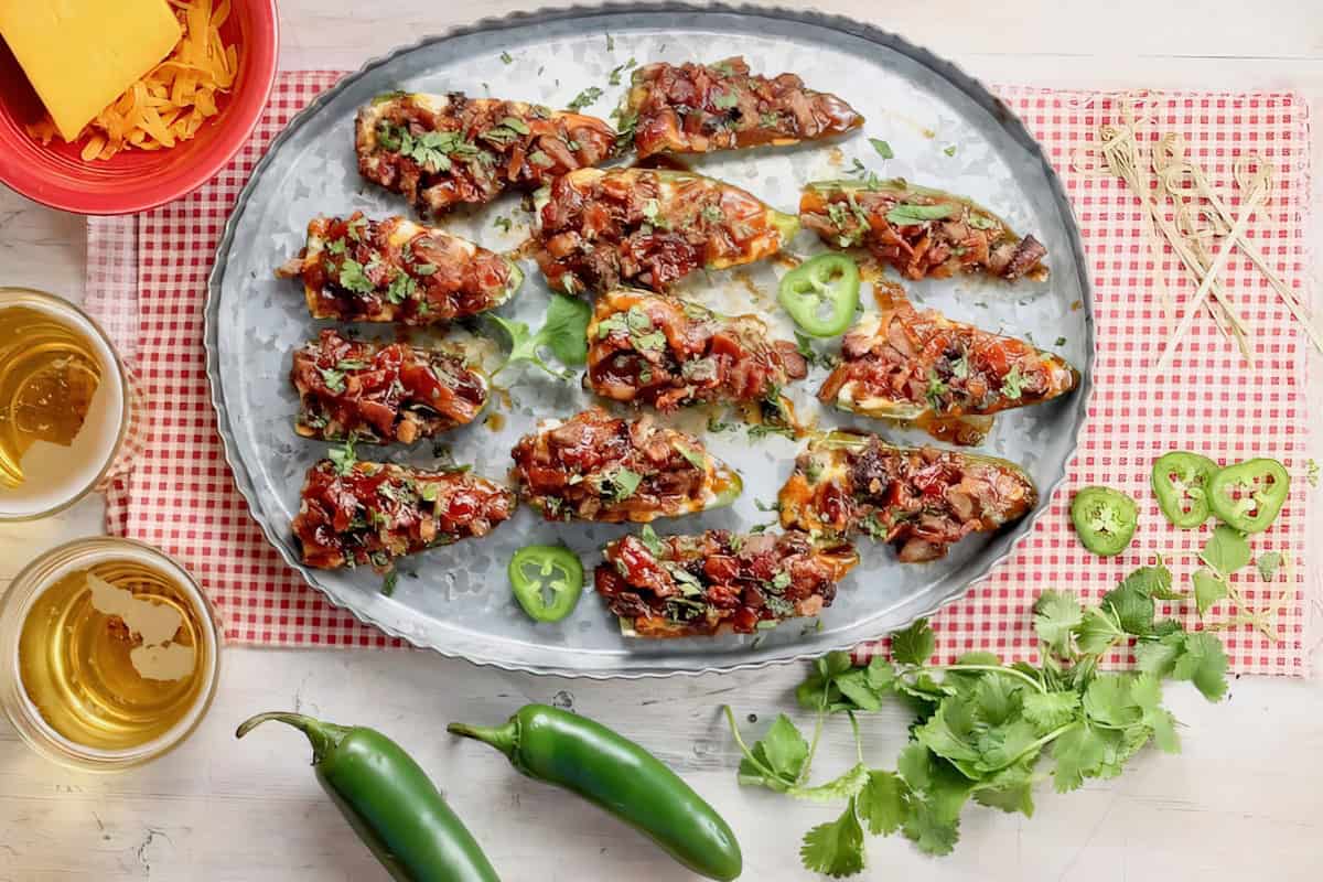 A tray of jalapeño poppers is topped with melted cheese and fresh herbs, set on a checkered cloth. Surrounding the tray are sliced jalapeños and cilantro. Two drinks are placed next to a bowl of shredded cheese.