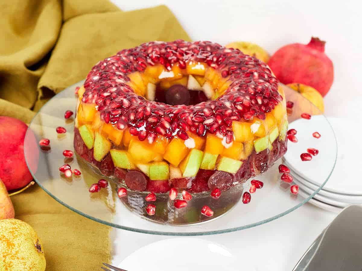 A colorful fruit jelly ring decorated with pomegranate seeds on a glass plate. The jelly is layered with grapes, melon, and mango pieces. Nearby are whole pomegranates, a pear, and an olive green cloth. Plates and utensils are in the background.