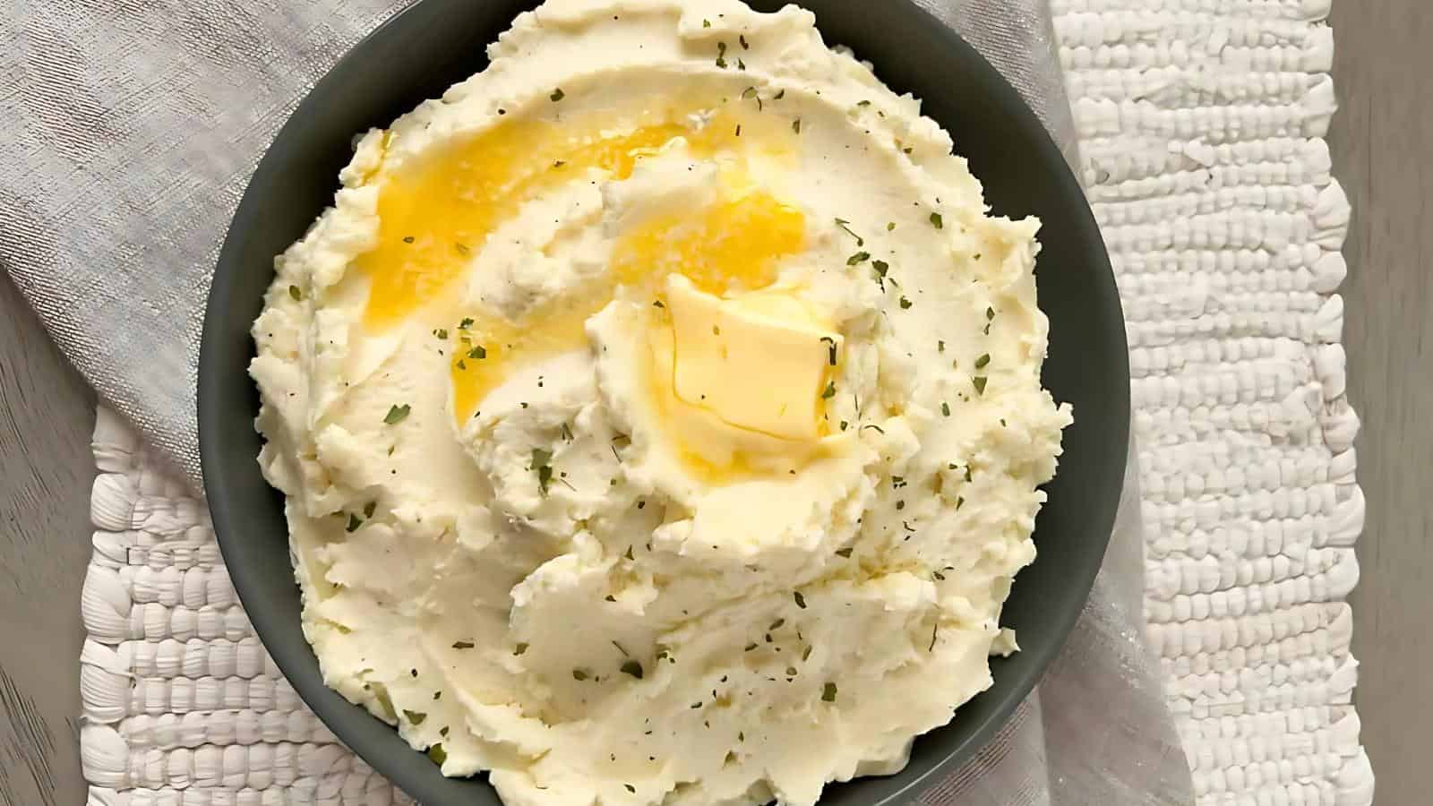 A bowl of mashed potatoes with a pat of butter on top, sprinkled with herbs, set on a woven cloth.