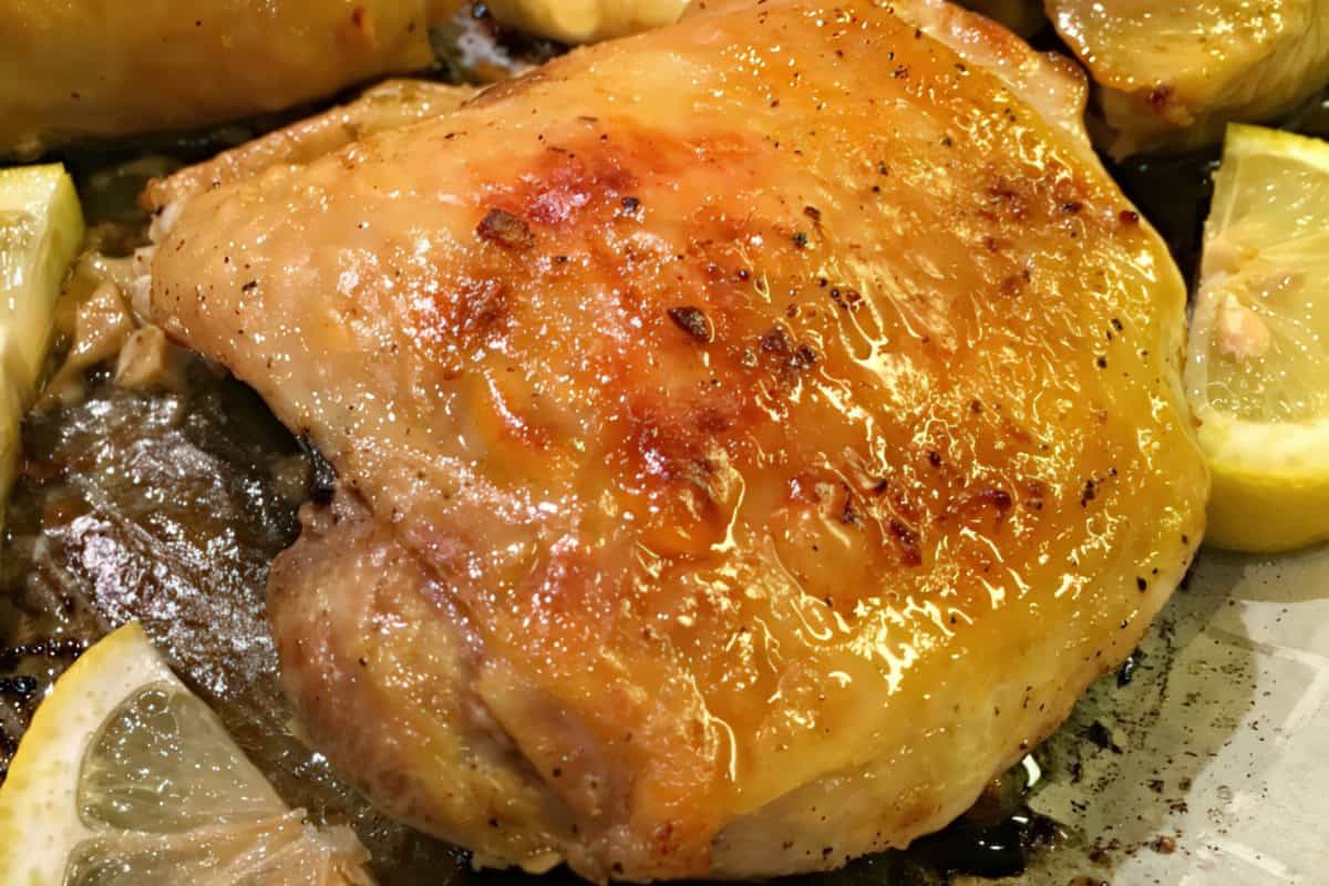 Close-up of a golden-brown, baked chicken thigh garnished with slices of lemon. The skin is crispy and seasoned, with visible juices, set on a roasting tray.