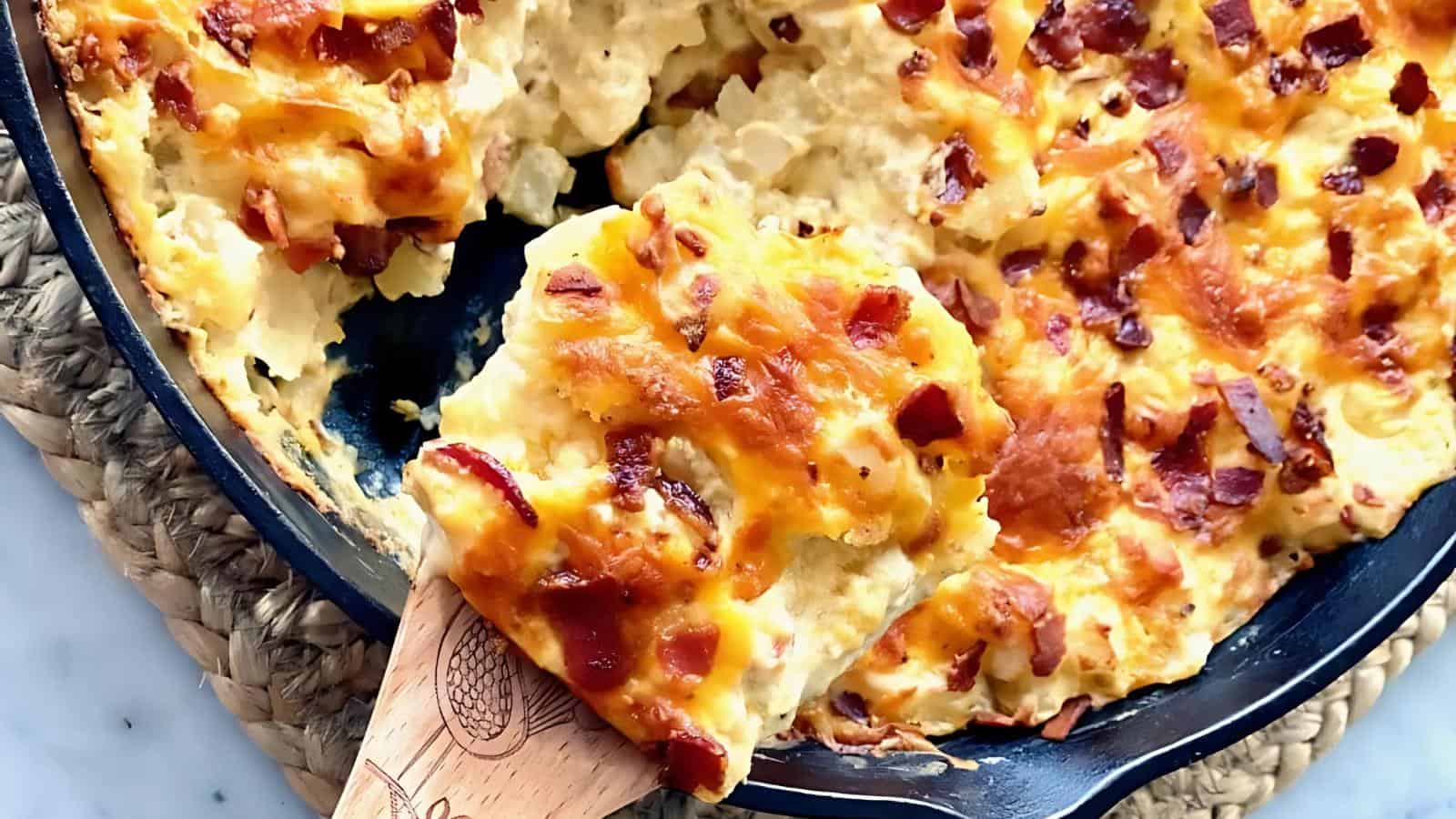 A skillet containing a cheesy baked dish with crispy bacon on top; a portion is being served with a wooden spoon.