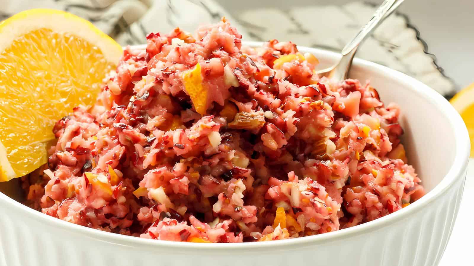 A bowl of cranberry relish with chunks of cranberries, oranges, and apples. A spoon rests in the bowl, and a slice of orange is placed on the rim. The relish is finely chopped, showcasing a vibrant mix of red and orange hues.