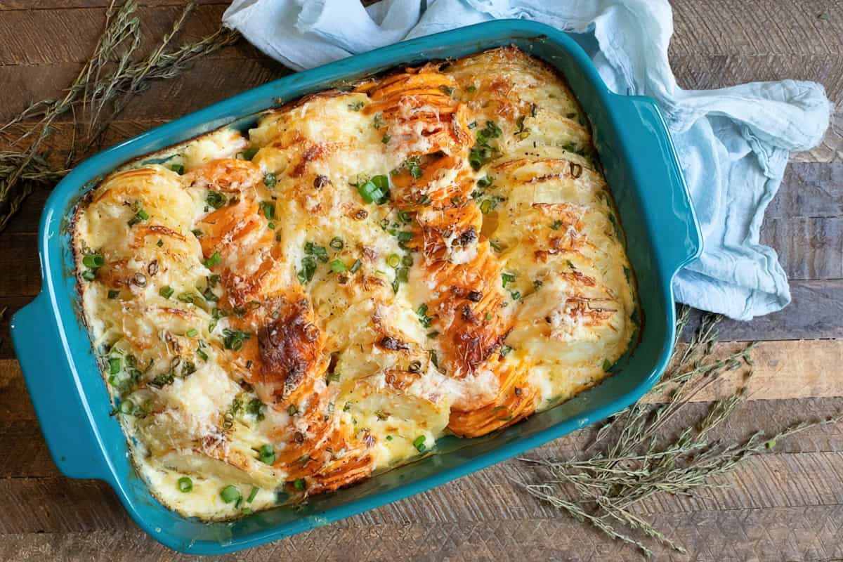 A blue dish containing baked potato gratin, topped with herbs and melted cheese, on a wooden table.