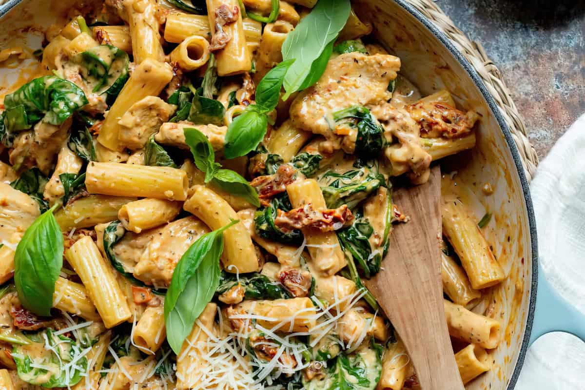 A skillet containing pasta mixed with spinach, sun-dried tomatoes, and basil, topped with shredded cheese. A wooden spoon rests in the skillet.