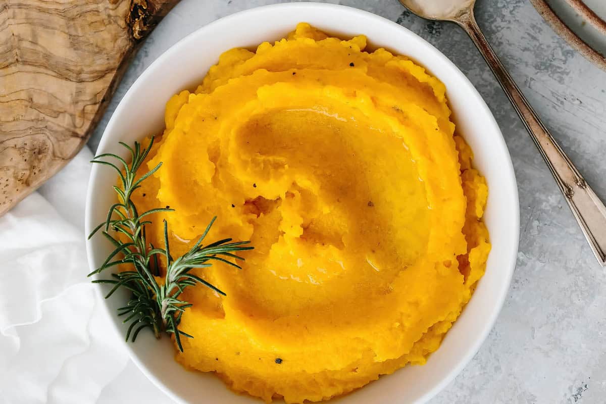 A bowl of smooth, creamy mashed butternut squash garnished with a sprig of rosemary. The bright yellow-orange mash is set on a gray surface, with a wooden spoon and a piece of rustic wood nearby.