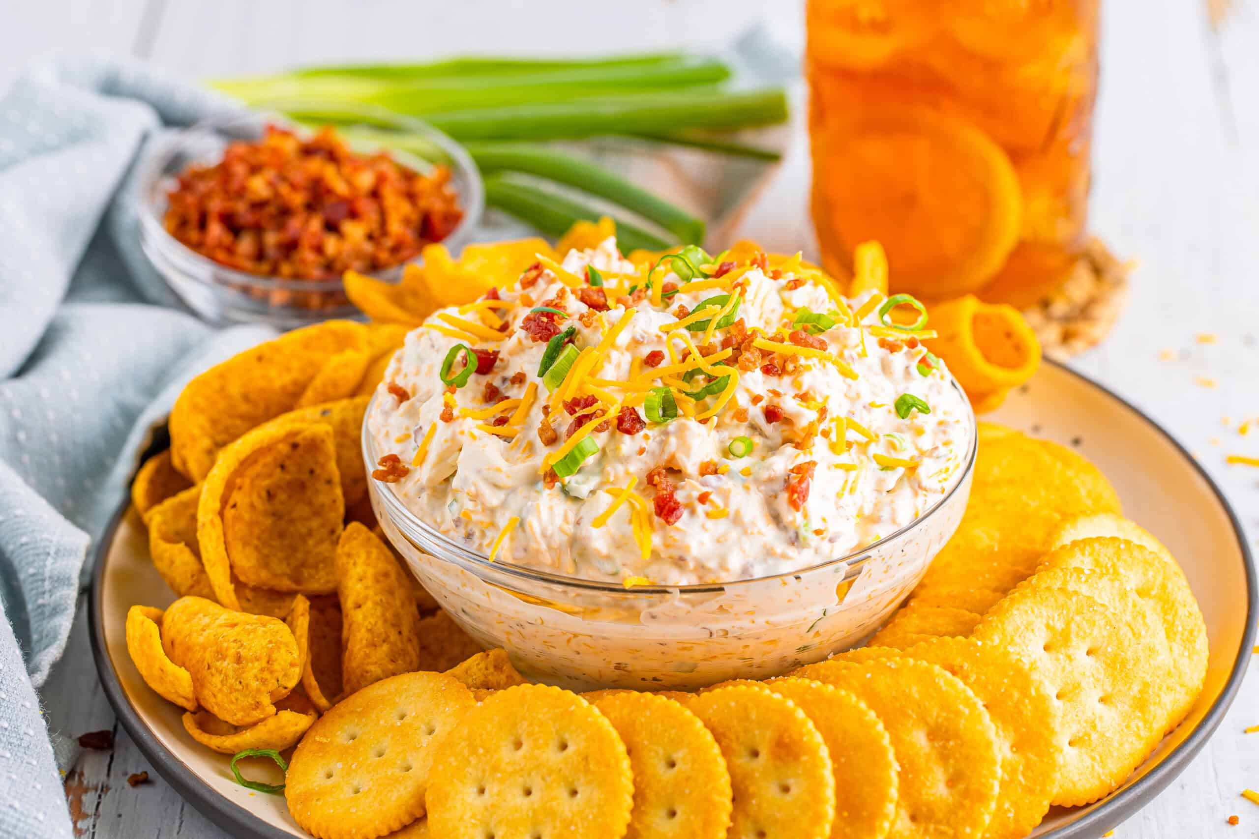 A bowl of dip topped with bacon and green onions is situated on a plate, which also holds crackers and corn chips. In the background, there is a bowl containing bacon bits and green onions.