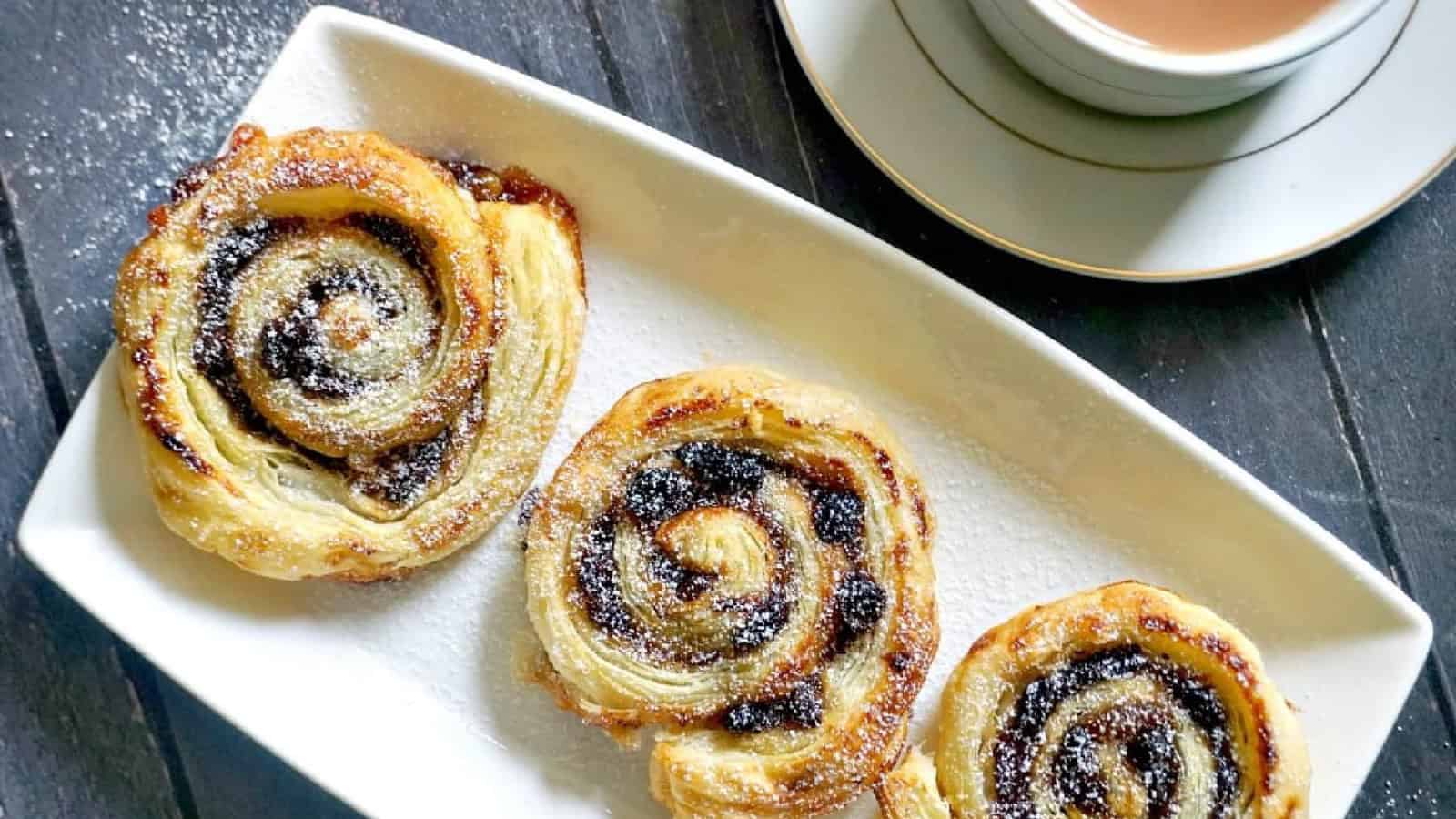 A rectangular white plate holding three spiral pastries sprinkled with powdered sugar is placed on a dark table. A cup of coffee in a white saucer is set beside the plate.