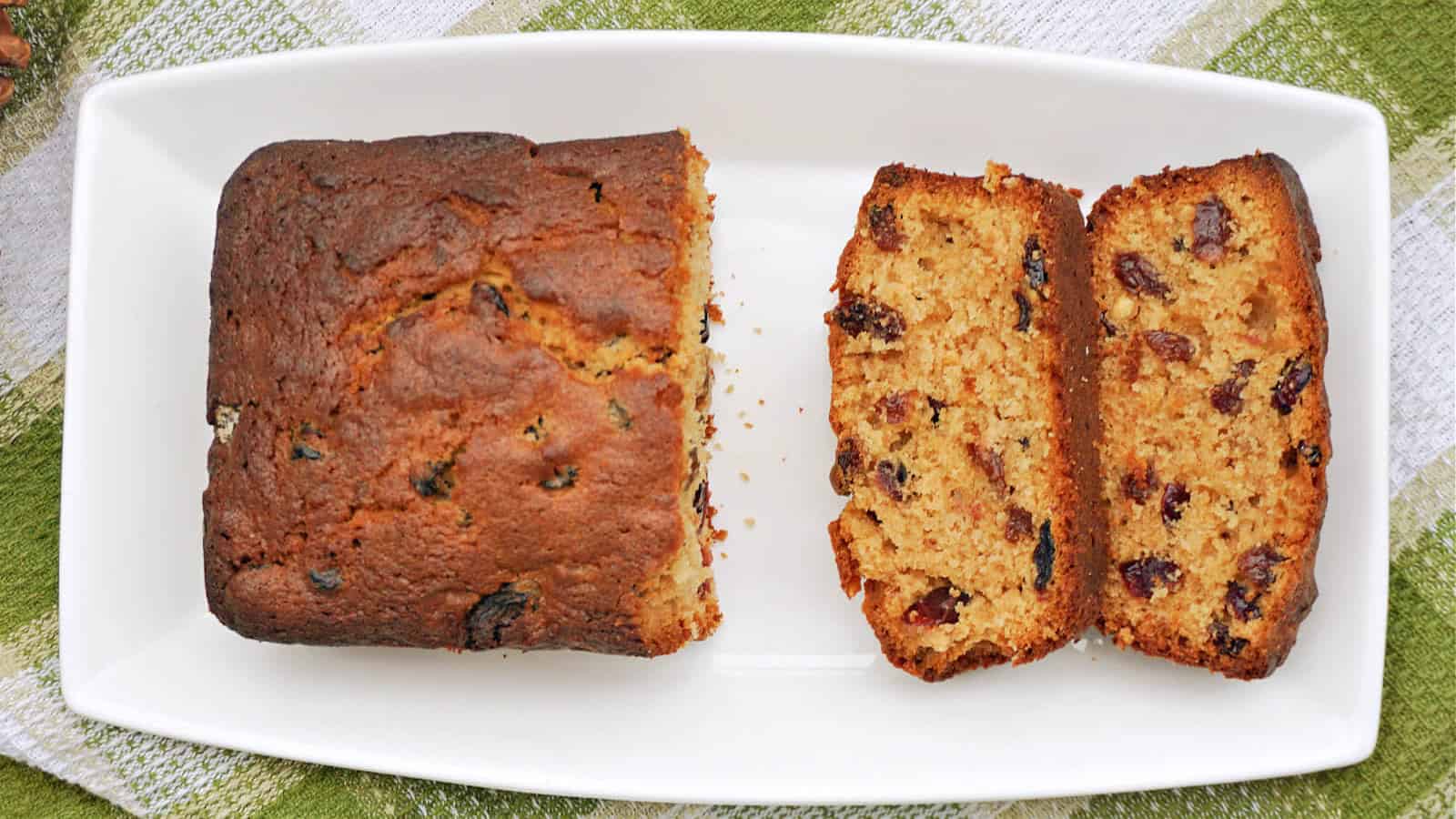 A loaf of fruitcake on a rectangular white platter with two slices cut. The cake appears to be homemade, featuring a golden-brown crust and dotted with raisins. The platter is set on a green and white checkered cloth.
