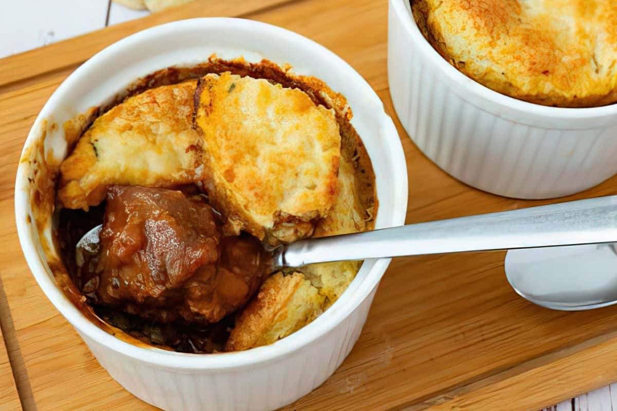 A small white ramekin filled with beef stew covered by golden brown, flaky pastry topping. A silver spoon rests on top, ready to scoop the dish. The ramekin is placed on a wooden surface.