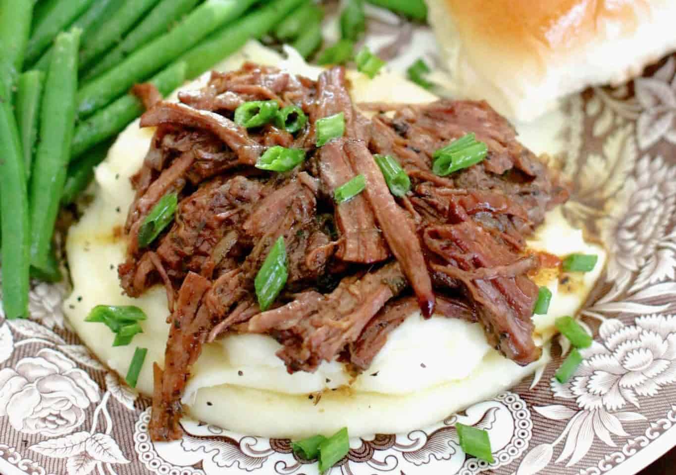 The image shows shredded beef on mashed potatoes, garnished with chopped green onions, accompanied by green beans and a roll on a decorative plate.