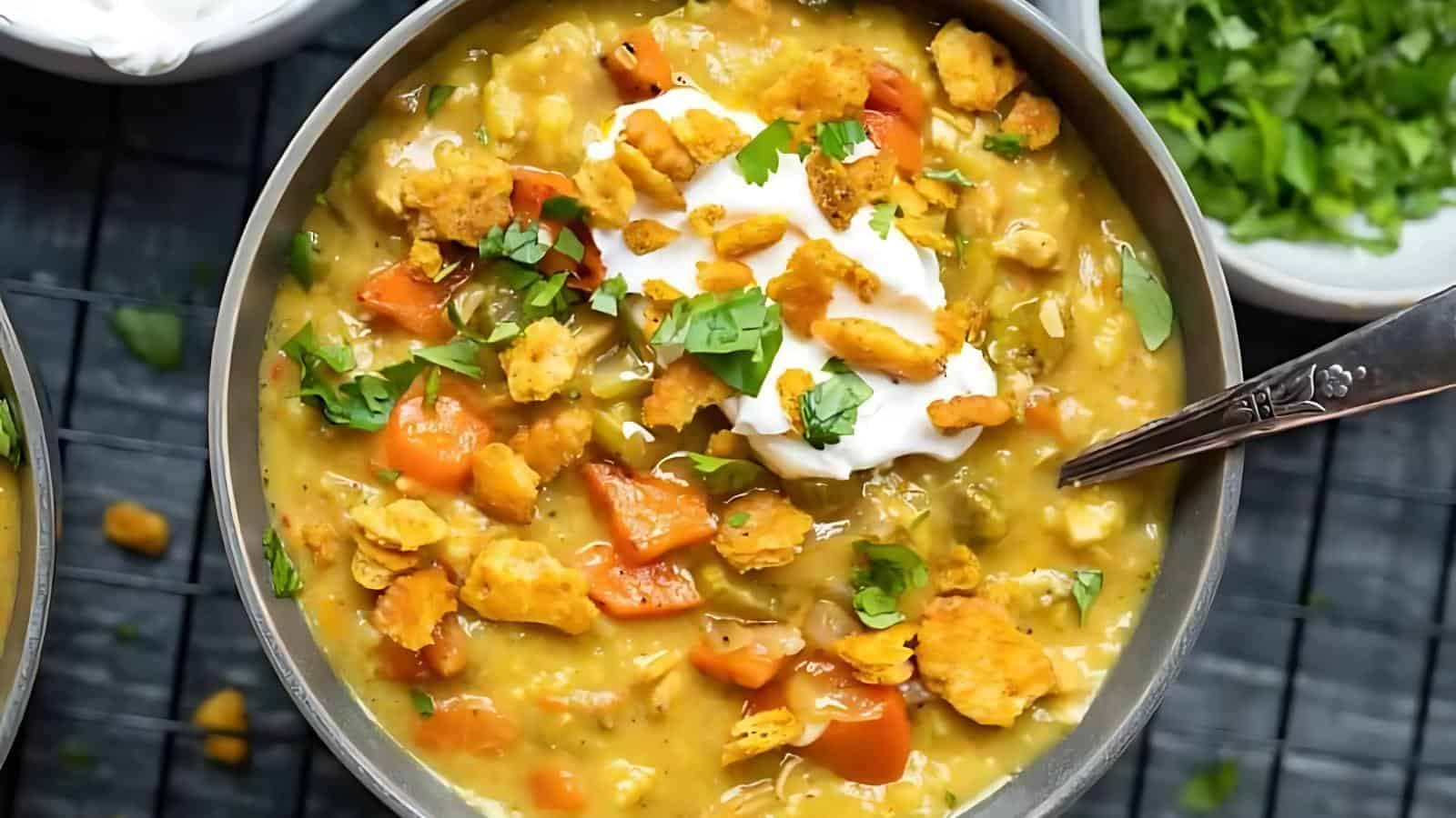 A bowl of vegetable soup topped with sour cream, crushed crackers, and chopped herbs, with a spoon inside. A small dish with herbs is in the background.