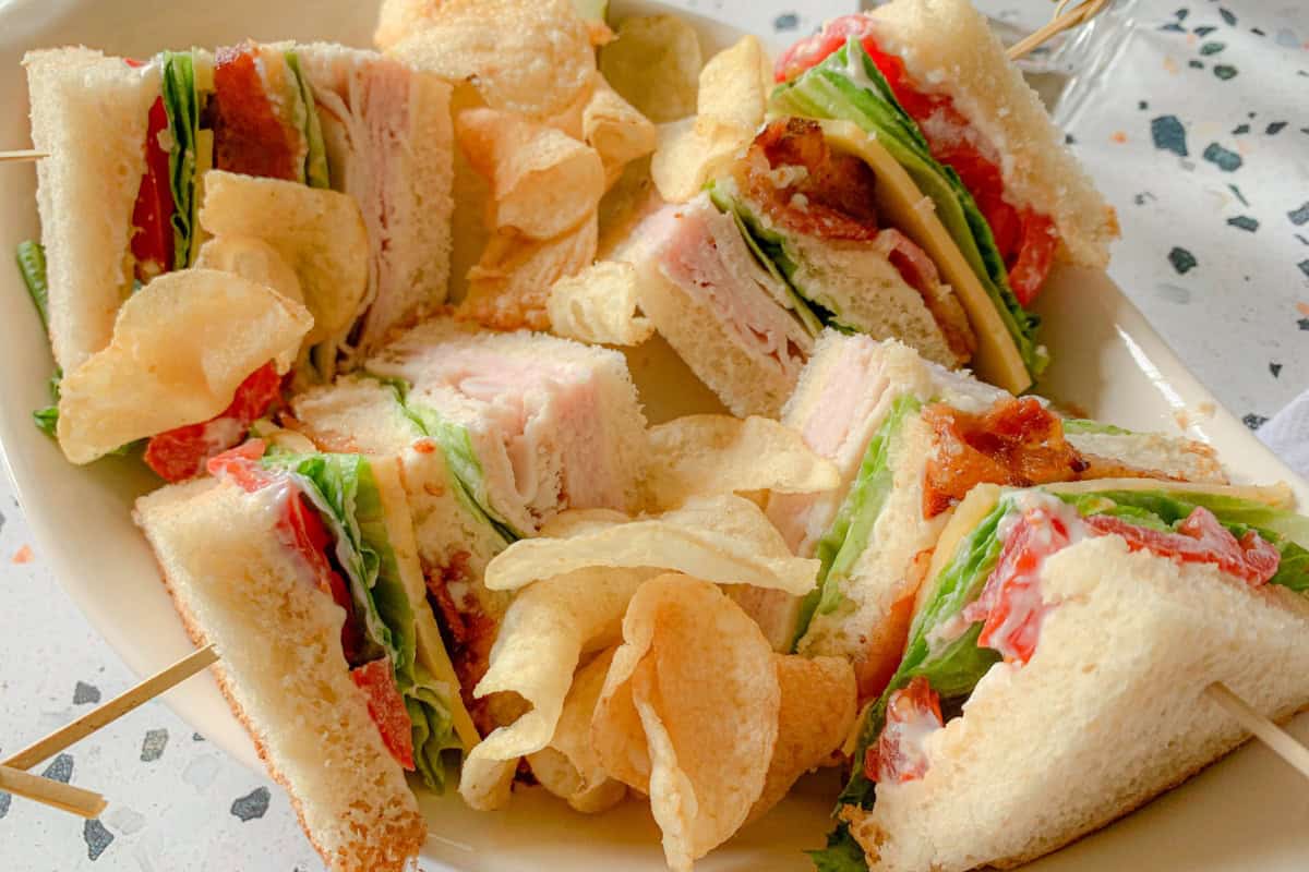 A plate of club sandwiches cut into quarters, with layers of turkey, bacon, lettuce, and tomato, garnished with potato chips. Skewers hold the sandwiches together, and the plate is set on a speckled countertop.