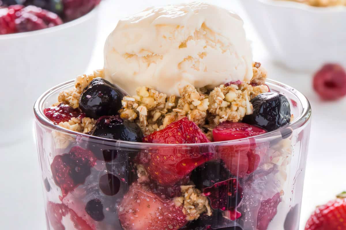 A glass filled with mixed berries and granola, topped with a scoop of creamy ice cream. The dessert is vibrant with red strawberries, blueberries, and blackberries, providing a refreshing and colorful presentation.