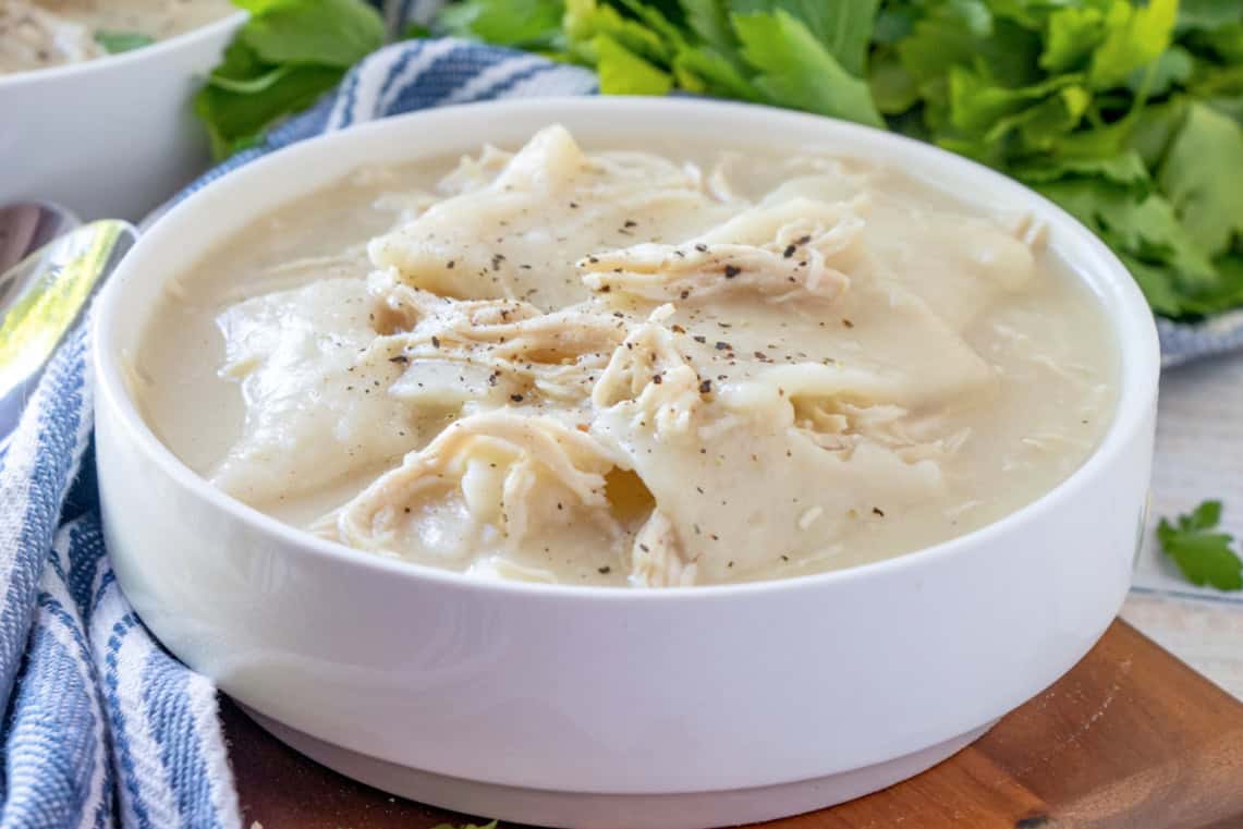 A white bowl filled with creamy chicken and dumplings, garnished with cracked black pepper, sits on a wooden surface beside a blue-striped cloth.