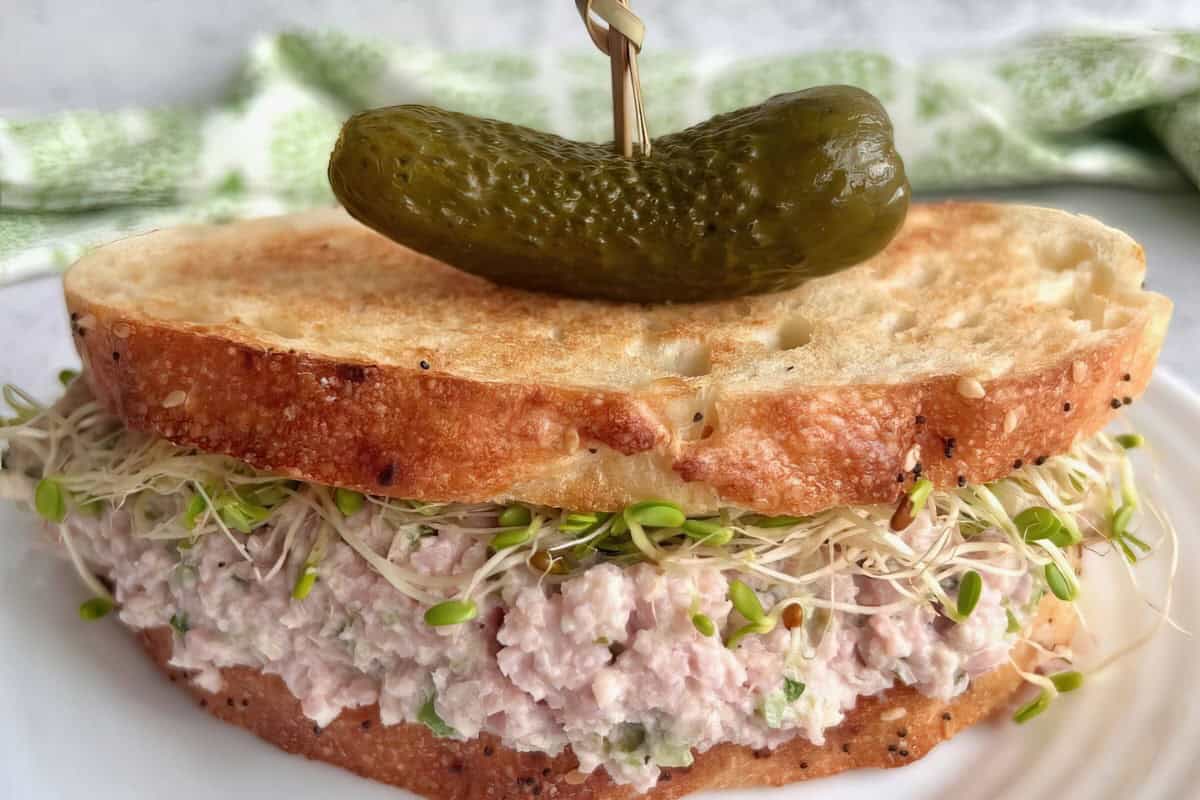 The image features a sandwich with toasted bread, filled with tuna salad and sprouts. It is topped with a small pickle secured by a toothpick.