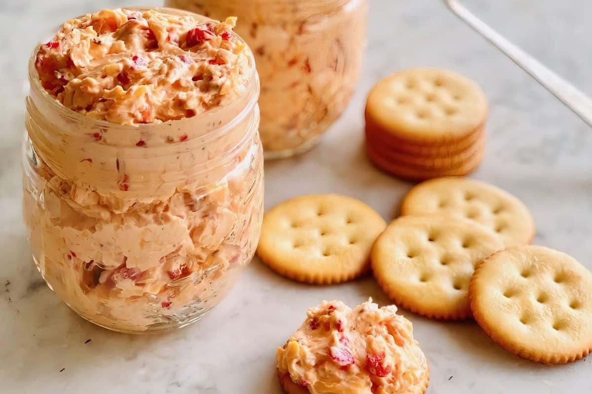 The image shows jars of pimento cheese spread on a marble surface, with round crackers nearby. One cracker has cheese spread on top.