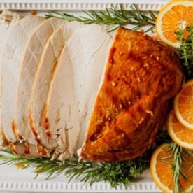 Sliced roasted turkey breast, garnished with rosemary, thyme, and orange slices, on a white platter.
