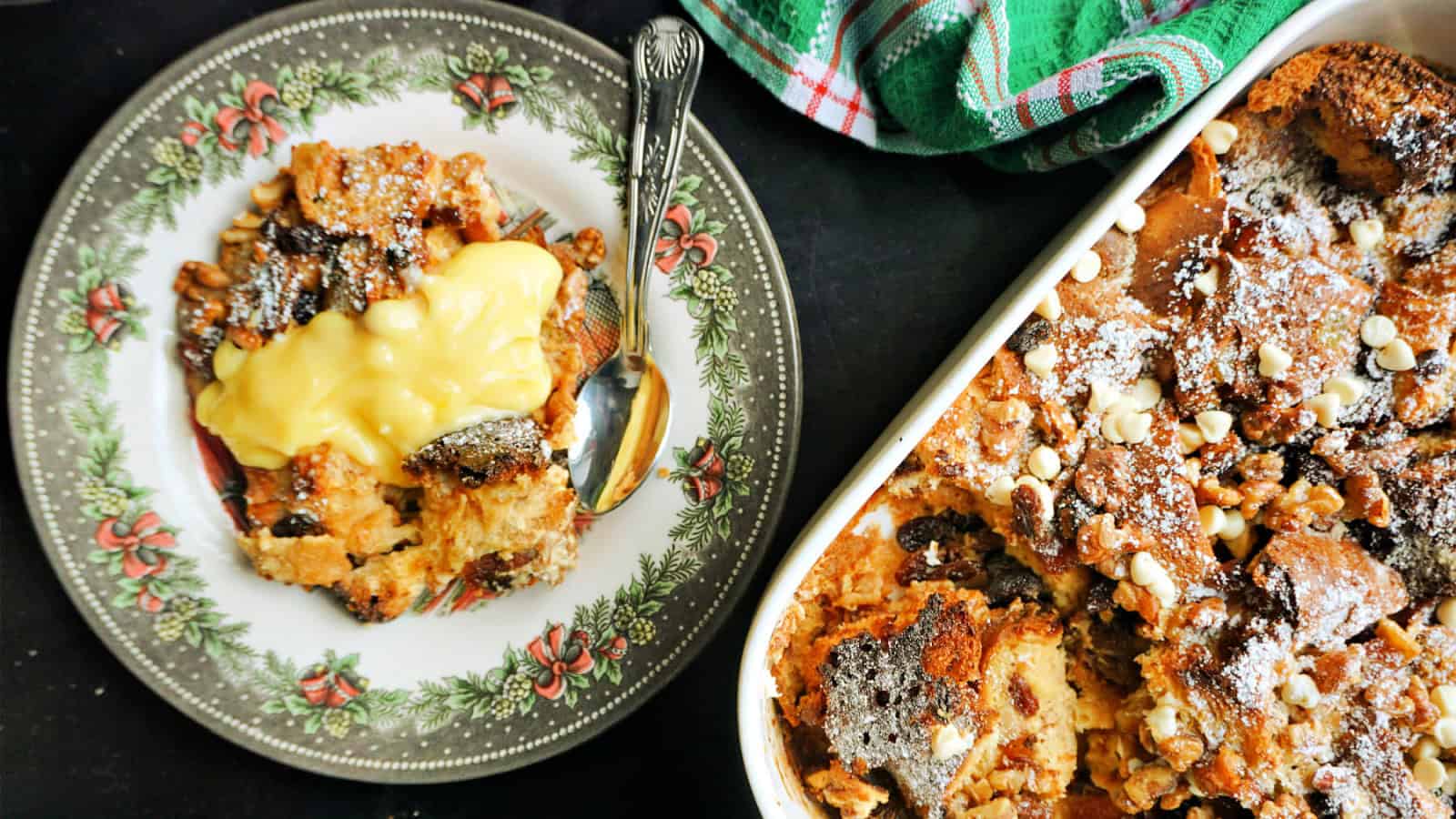 A plate with a serving of chocolate bread pudding topped with vanilla sauce next to a baking dish filled with more pudding. A green kitchen towel and a spoon are on the side.